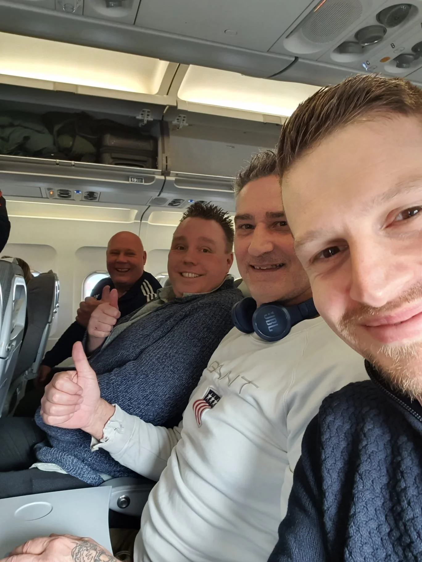 Four men sitting in airplane seats, smiling and giving thumbs up.