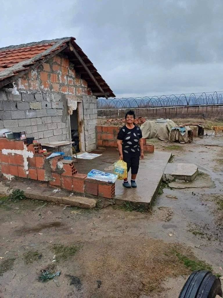 A young boy standing on a small concrete porch in front of a partially constructed brick and concrete house, holding a plastic bag with purchases, with farm equipment and a dog in the background under a cloudy sky.
