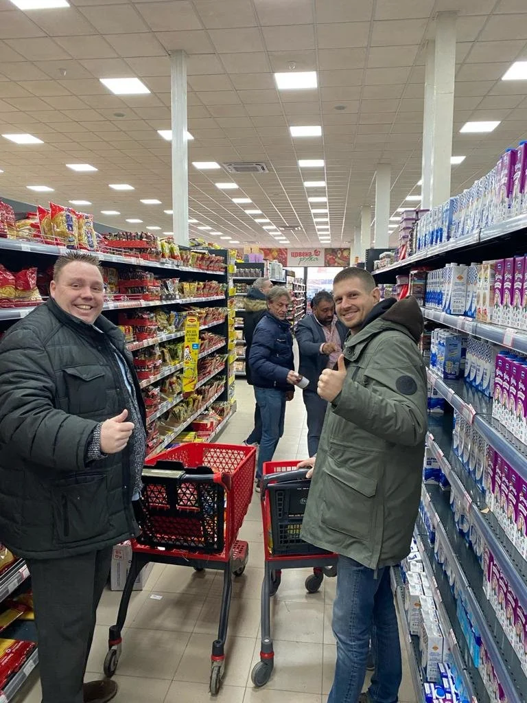 Four people in a grocery store aisle with shopping carts, smiling and giving thumbs up. Two men in the foreground, one in a black jacket and the other in a green jacket, and two women in the background, all shopping.