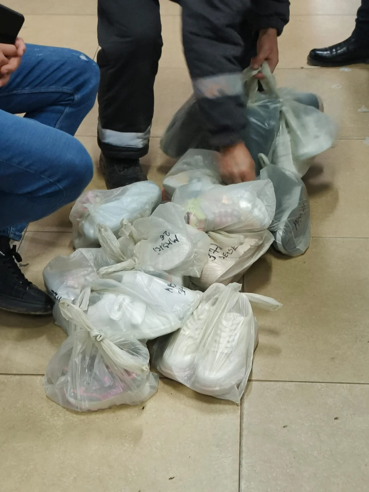 Multiple clear plastic bags filled with food, placed on a tiled floor, with people kneeling around them.