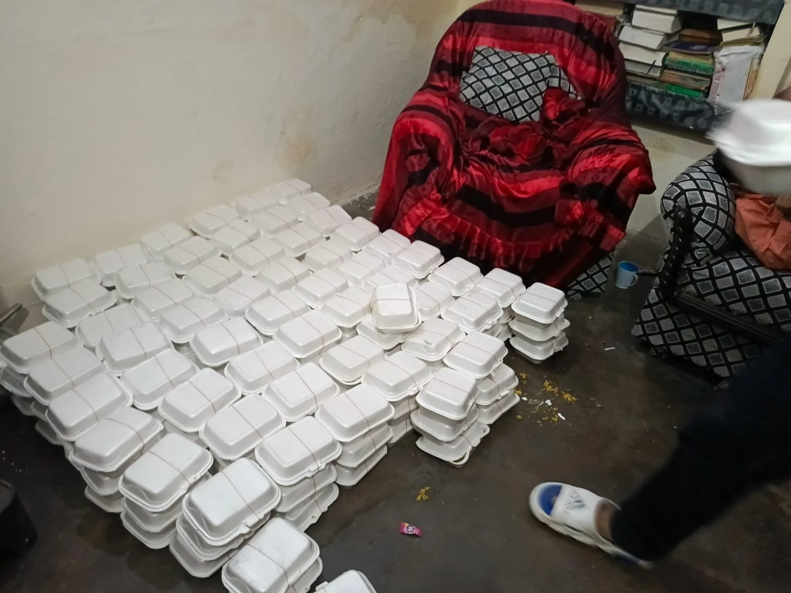 A large number of stacked white disposable food containers on the floor next to two patterned armchairs, one covered with a red blanket, in a room with a cluttered bookshelf.