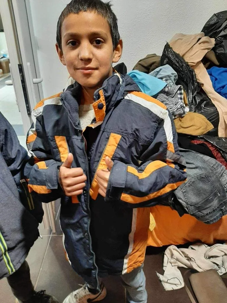 A young boy giving two thumbs up, wearing a dark jacket with orange and white accents, standing in front of a table piled with clothes.
