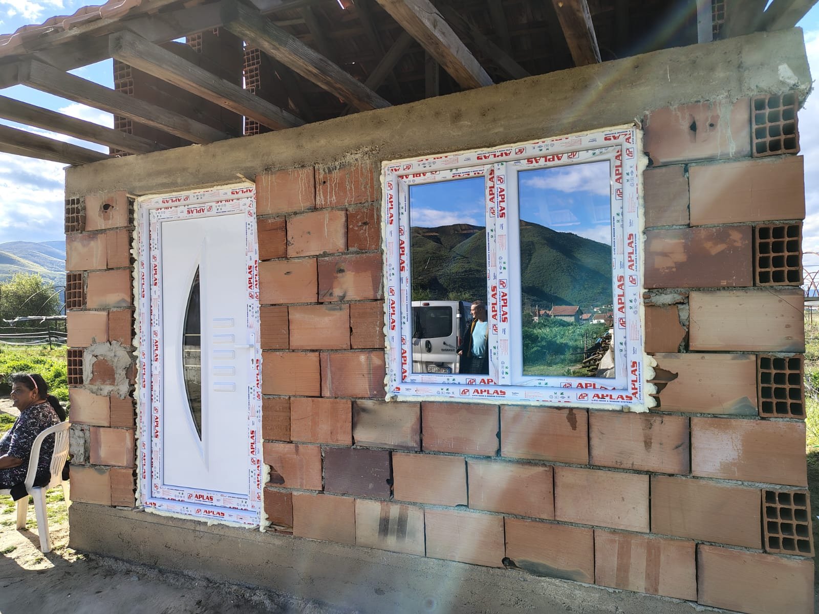 A brick house under construction featuring an exterior door and window frame with reflective glass, set against a scenic mountainous landscape. A woman sits outside on a white chair, and a man is reflected in the window, with a truck visible in the background.