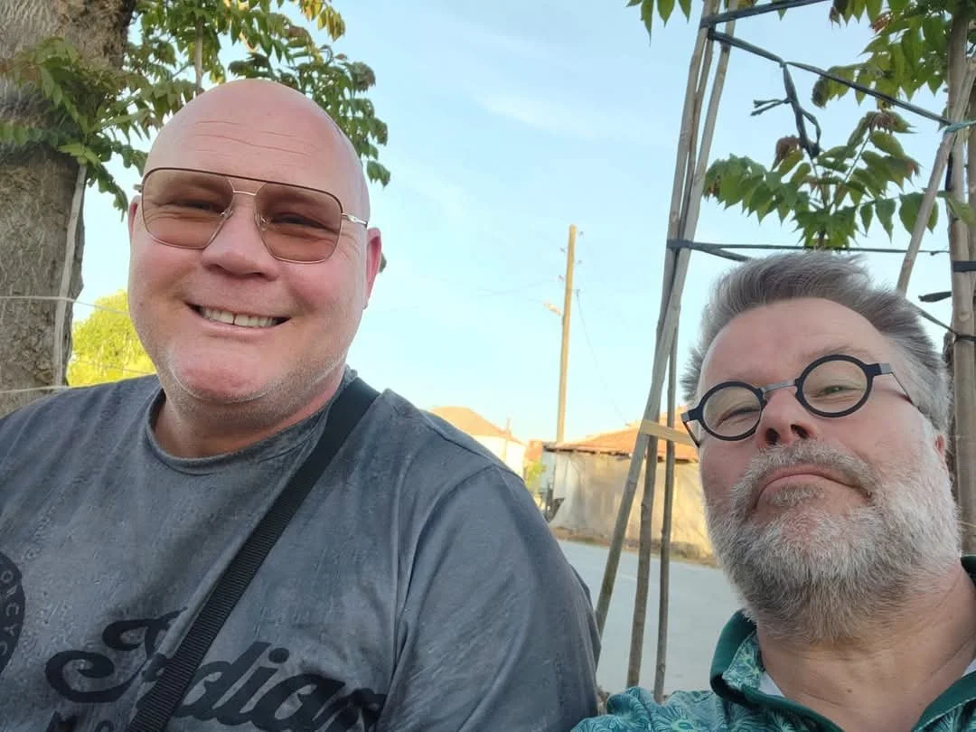 Two smiling middle-aged men with glasses outdoors on a sunny day, one with a bald head and the other with gray hair and beard, standing near a wooden structure and trees.