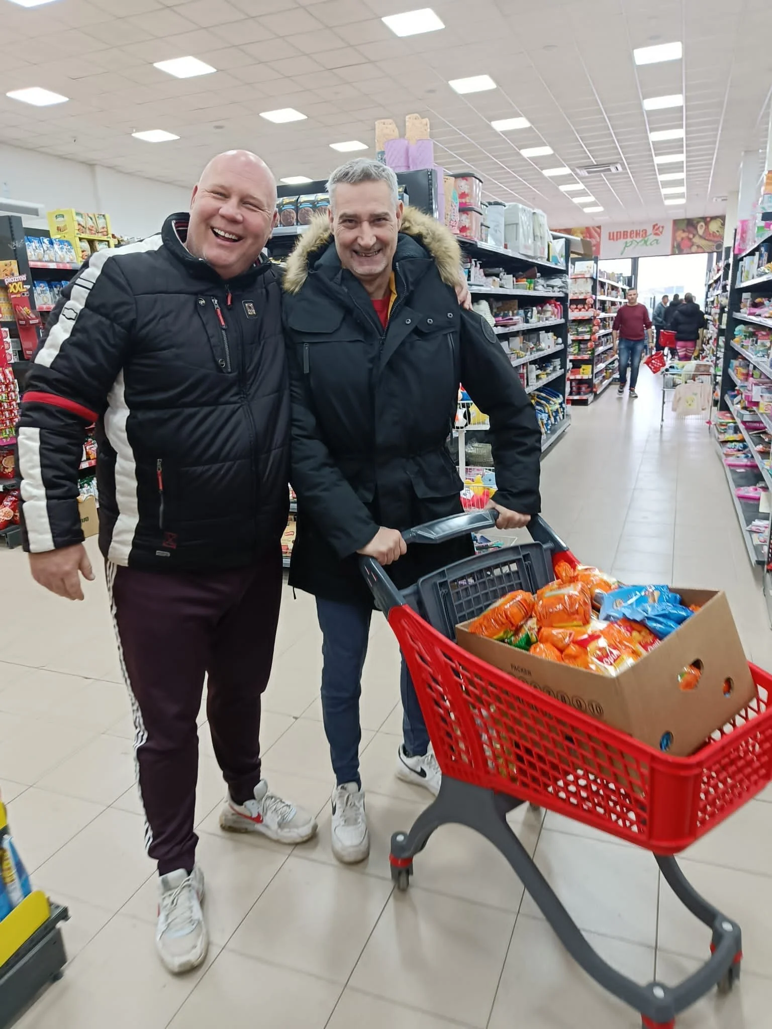 Two smiling men standing inside a grocery store, with one man pushing a shopping cart filled with snacks, surrounded by shelves filled with various products.