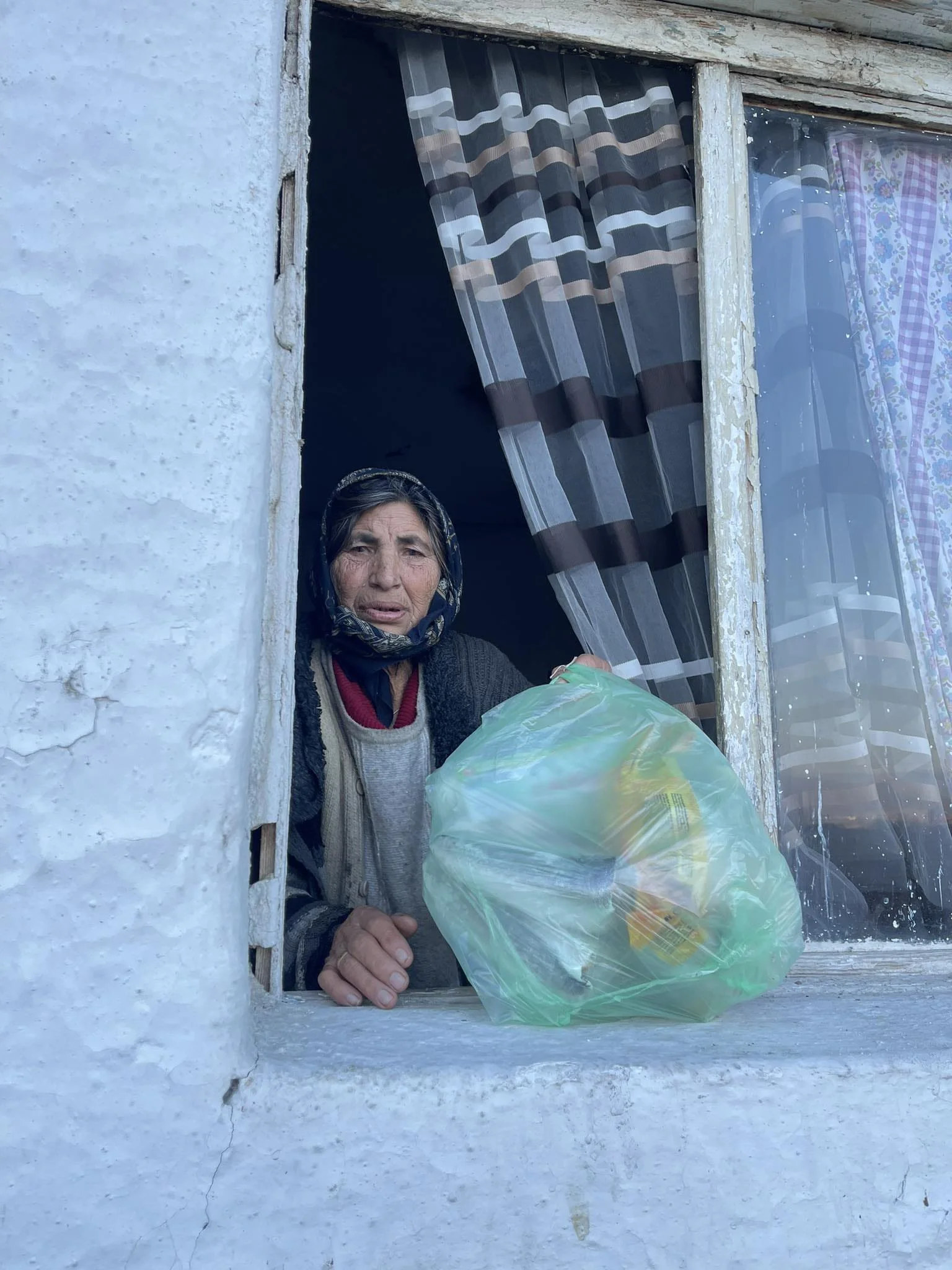 An elderly woman looks out from a window, holding a plastic bag with groceries outside her rustic home.