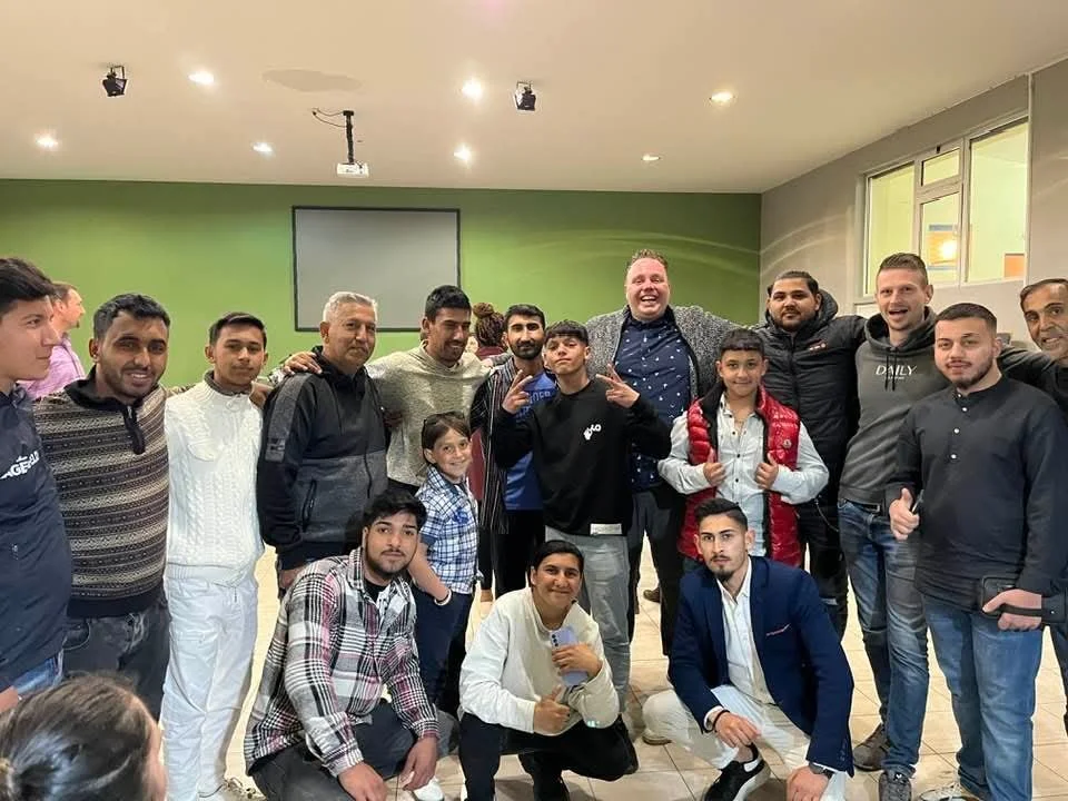 Group of people standing and kneeling indoors, smiling and posing for a photo. The background includes a green wall and a window.