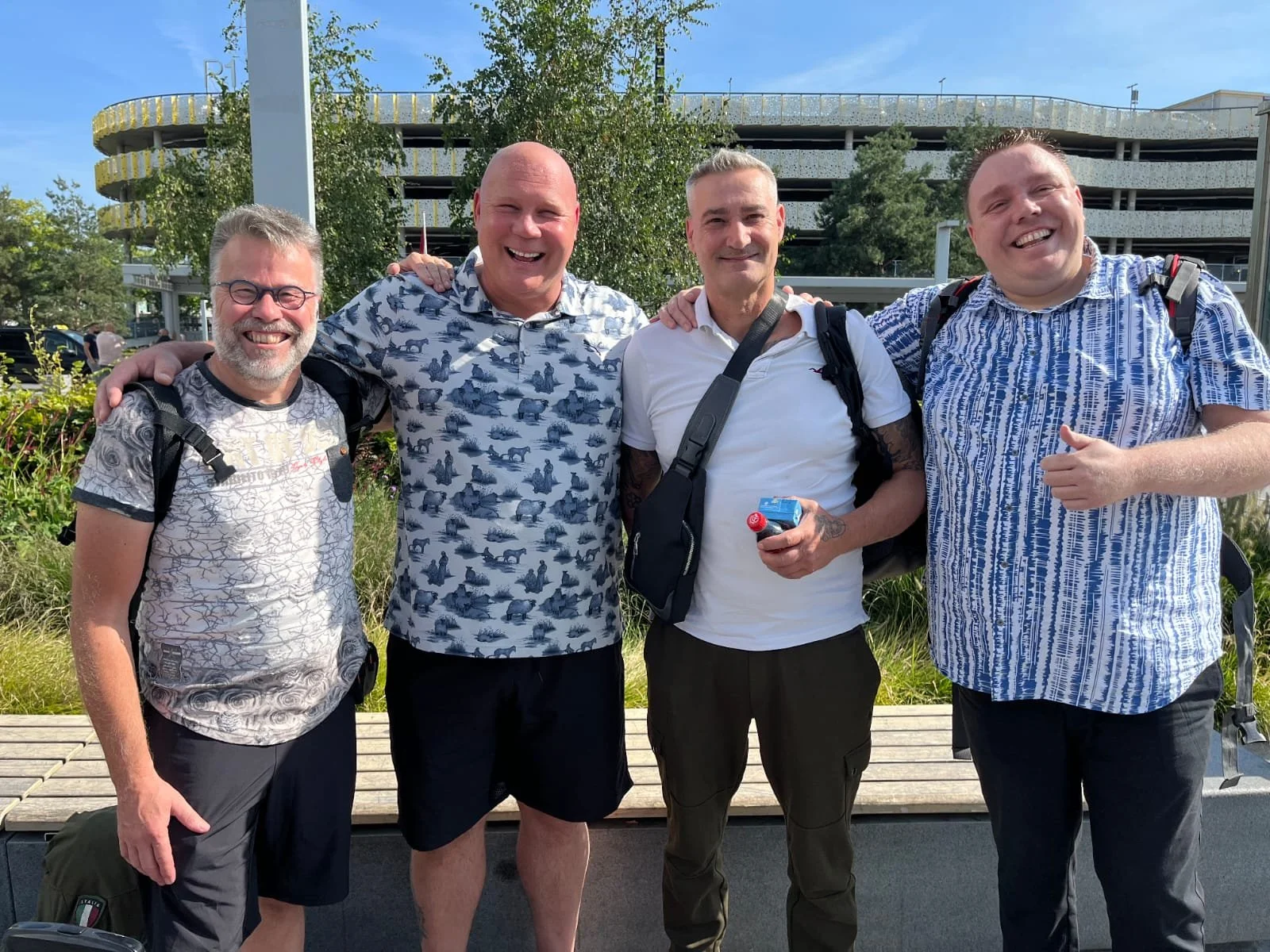 Four smiling men standing outdoors with trees and a building in the background, wearing casual shirts and backpacks.