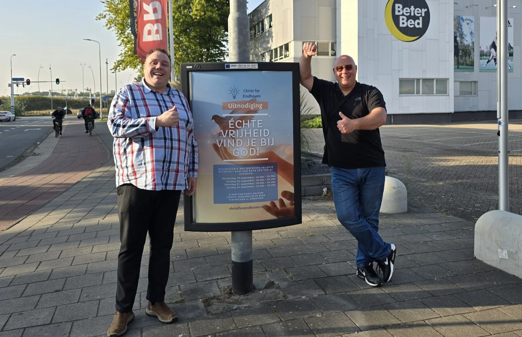 Two men standing on a sidewalk next to a digital sign with Dutch text, giving thumbs-up gestures. One man is dressed in a checkered shirt and dark pants, and the other in a black shirt and jeans. They are smiling, with a street and building in the background.