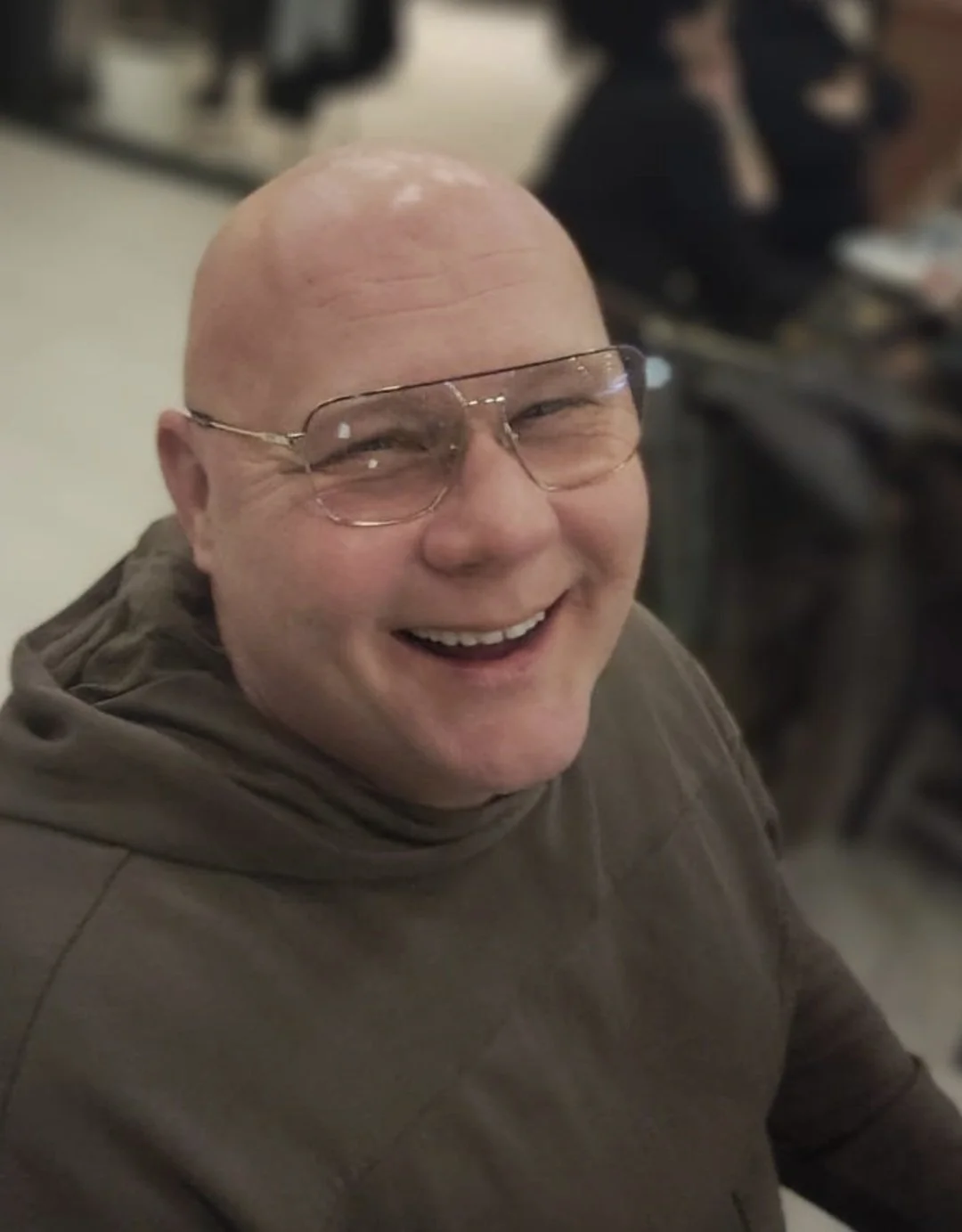 A bald man with glasses smiling widely, wearing a brown hoodie in an indoor setting.