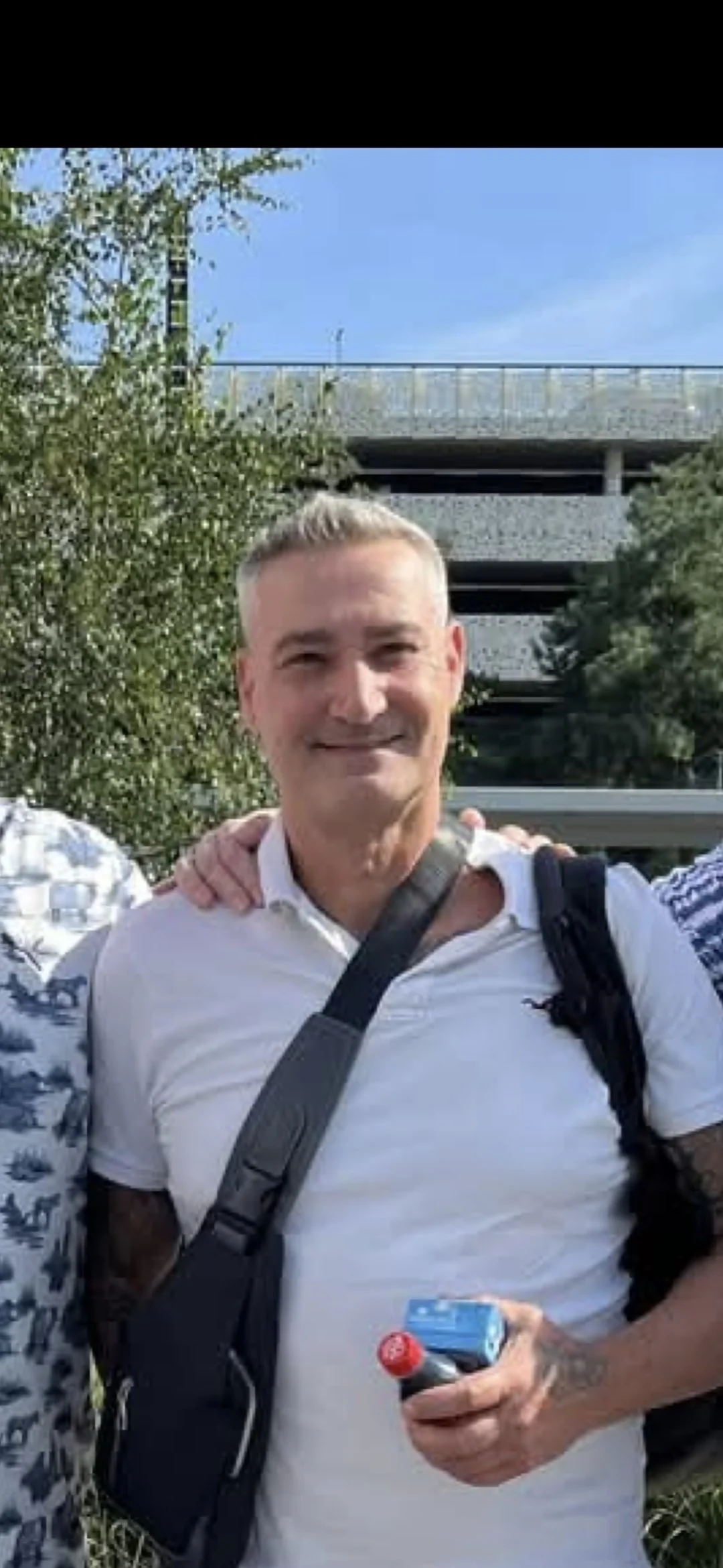 A smiling man with short gray hair wearing a white polo shirt, carrying a black shoulder bag, holding a small red-capped bottle and a blue pack, standing outdoors with trees and a multi-level parking garage in the background.