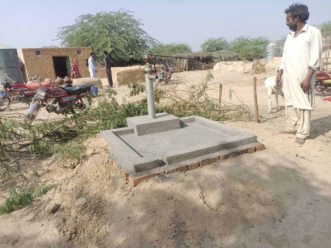 A newly installed hand pump water well in a rural village, with a man standing nearby and several motorcycles and villagers in the background.