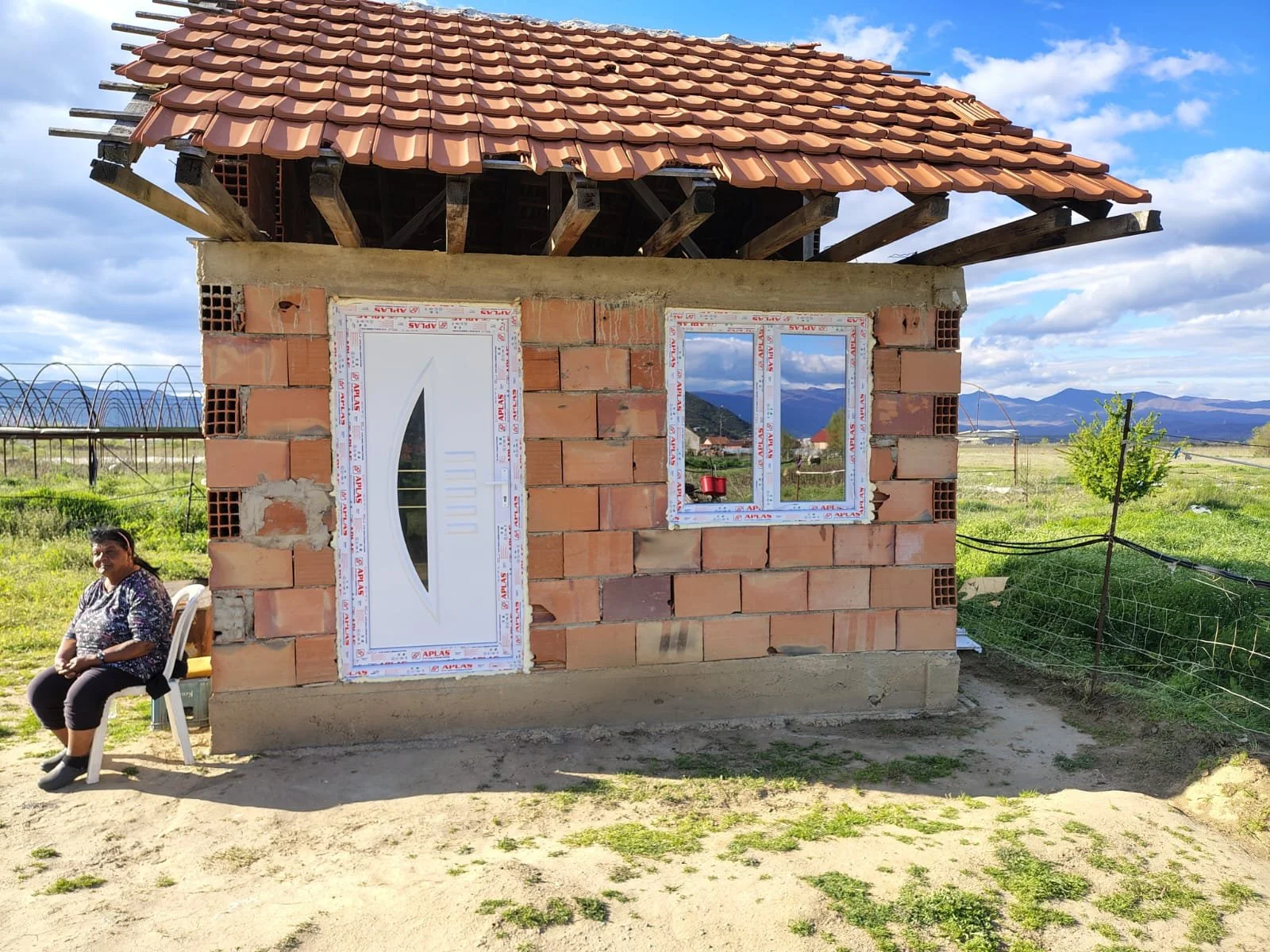 A small brick house under construction with a woman sitting on a white plastic chair beside it. The house has a tiled roof, a white door, and window frames. The scene is set outdoors with green fields, a tree, and mountain ranges in the background.