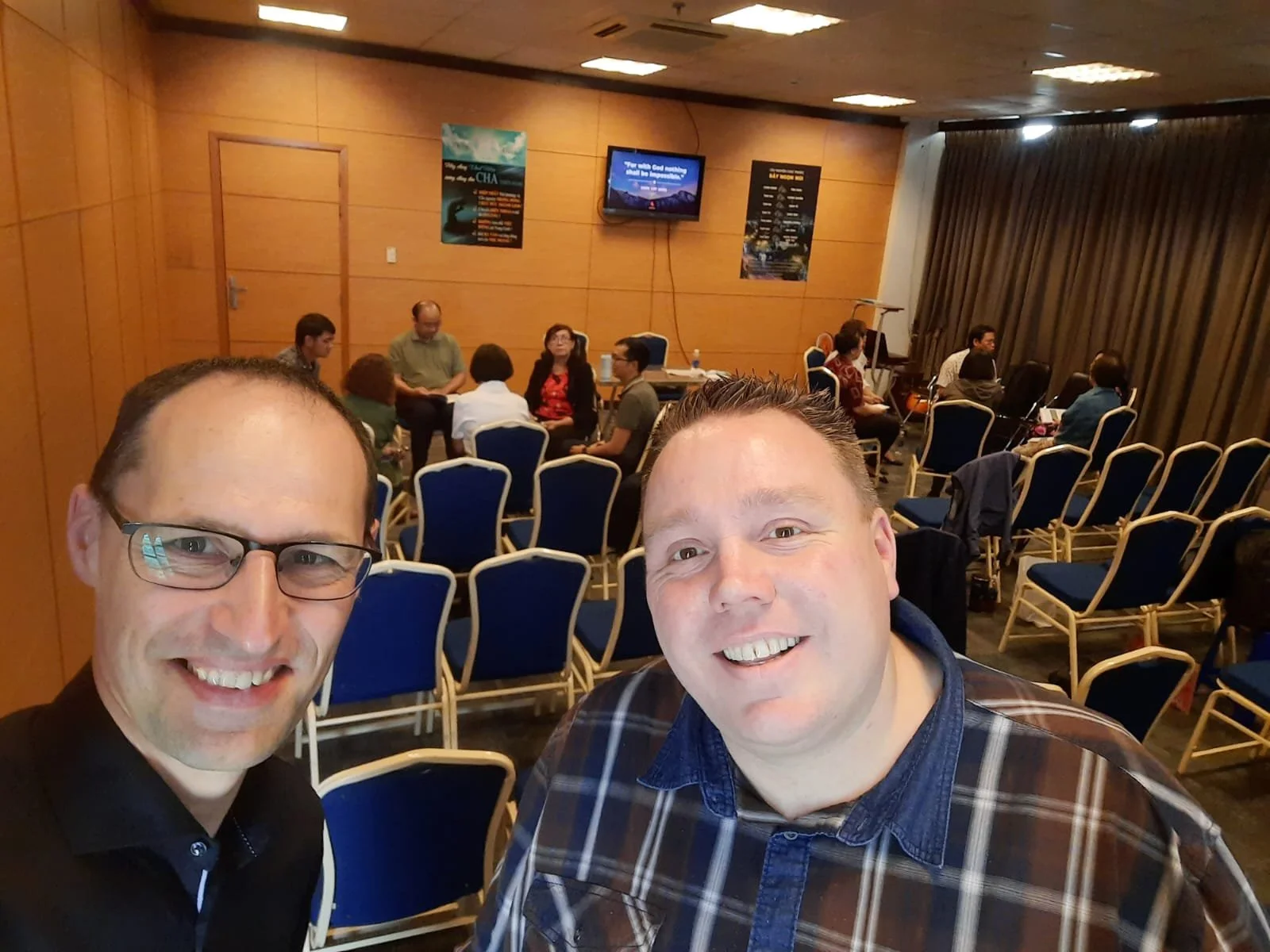 Two smiling men taking a selfie in a conference room with empty chairs and a group of people sitting in a circle in the background.
