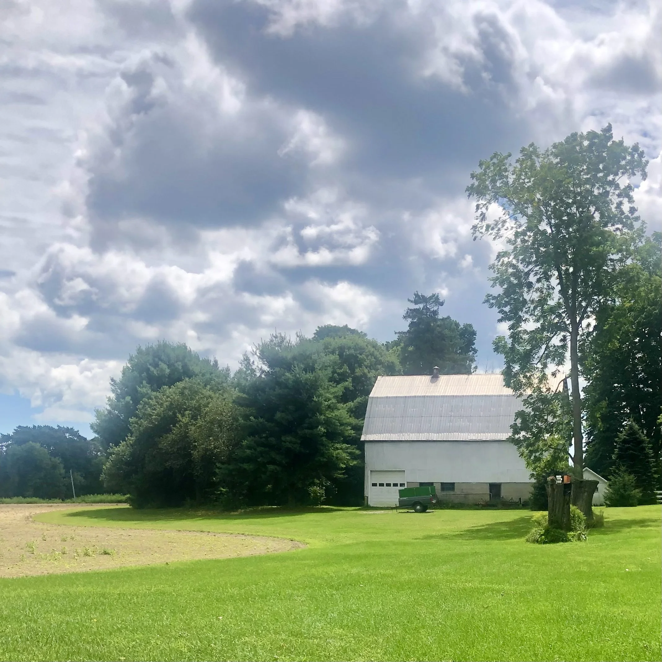 A barn with a white roof sits beside a grassy field under a cloudy sky. There are trees surrounding the barn, and a tractor is parked nearby.