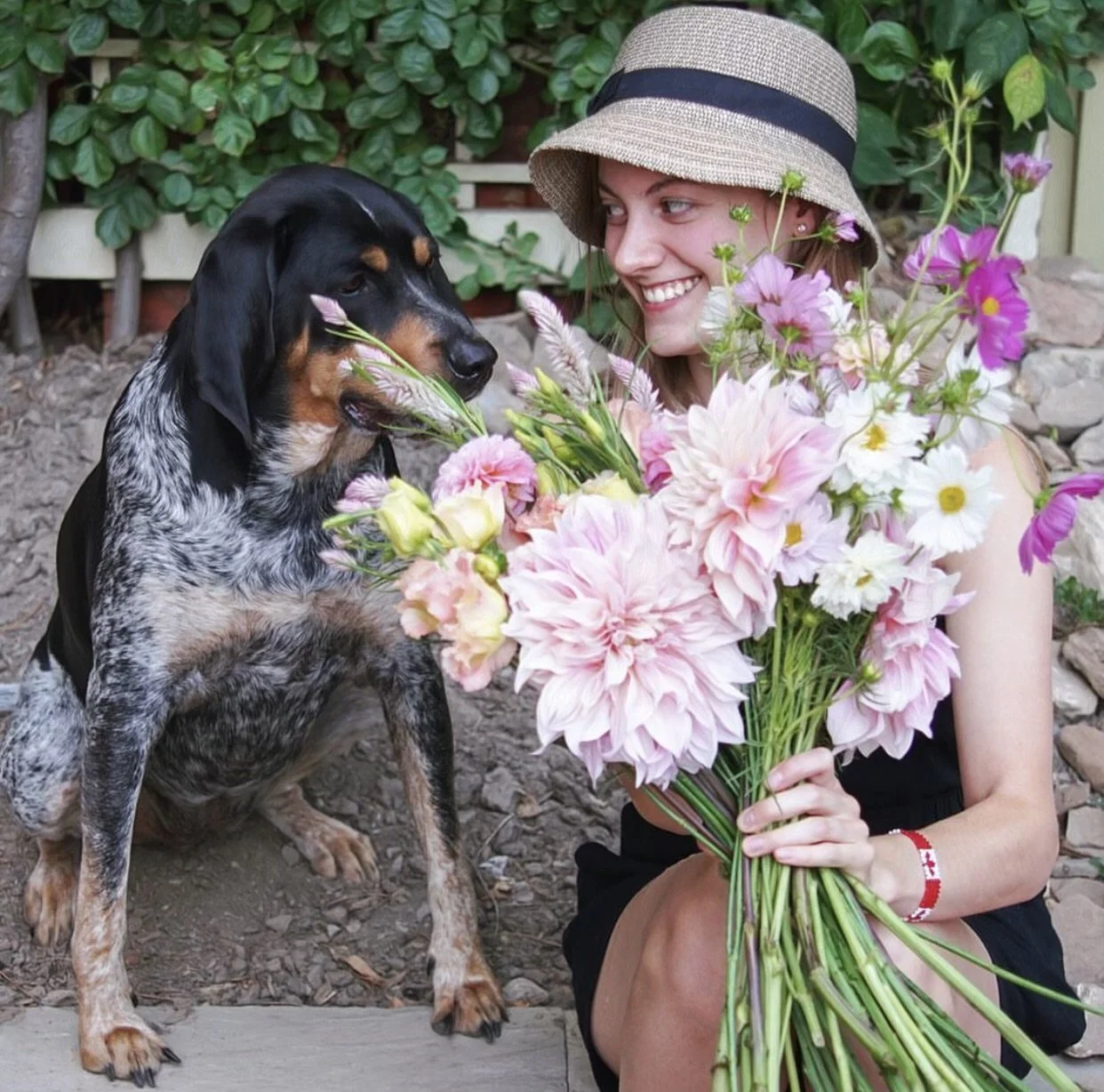 A person wearing a hat smiles at a dog while holding a large bouquet of pink and white flowers outdoors.
