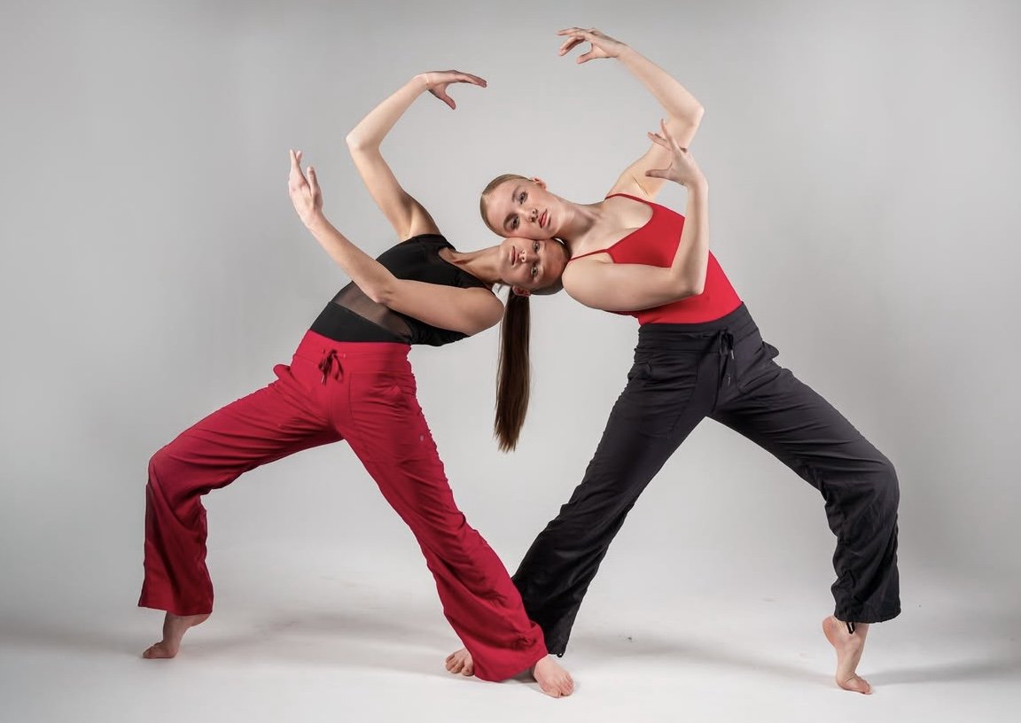 Two women in dance poses leaning their heads together, wearing athletic clothing, against a plain gray background.