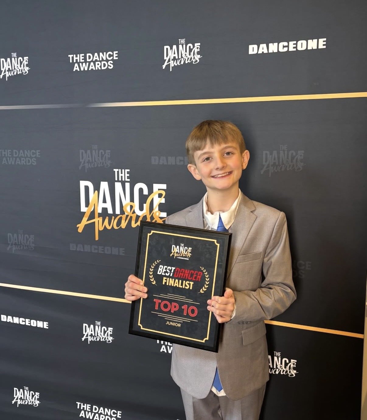 A young boy in a gray suit and tie holding a framed award plaque that reads 'Best Dancer Finalist Top 10 Junior', standing in front of a backdrop with 'The Dance Awards' logos.