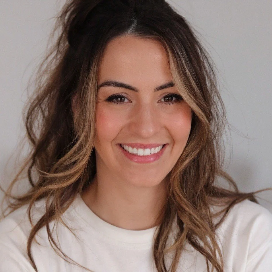 Woman with long brown hair smiling, wearing a black top and jeans, sitting indoors with a contemplative pose.