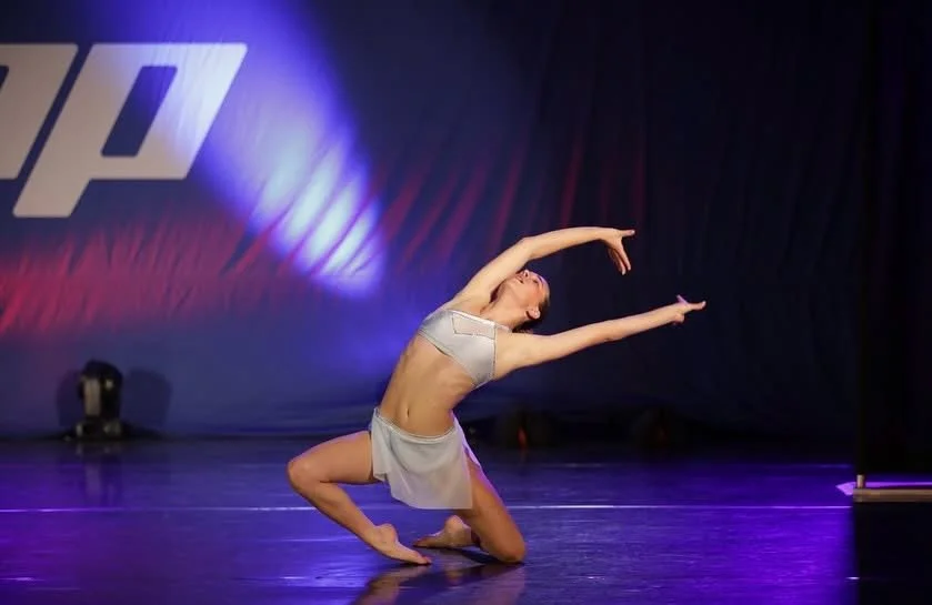 A female dancer performing on stage in a silver costume, kneeling with arms extended gracefully, with stage lighting and a dark backdrop.