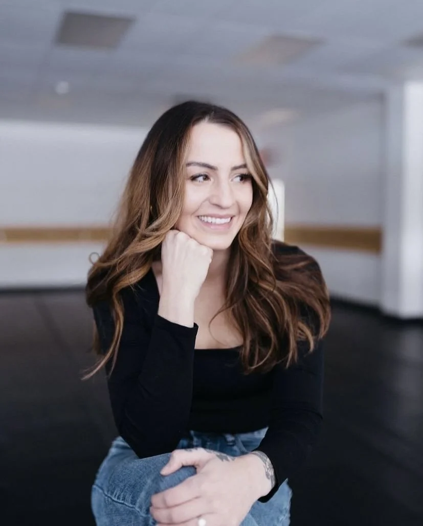 Woman with long brown hair smiling, wearing a black top and jeans, sitting indoors with a contemplative pose.