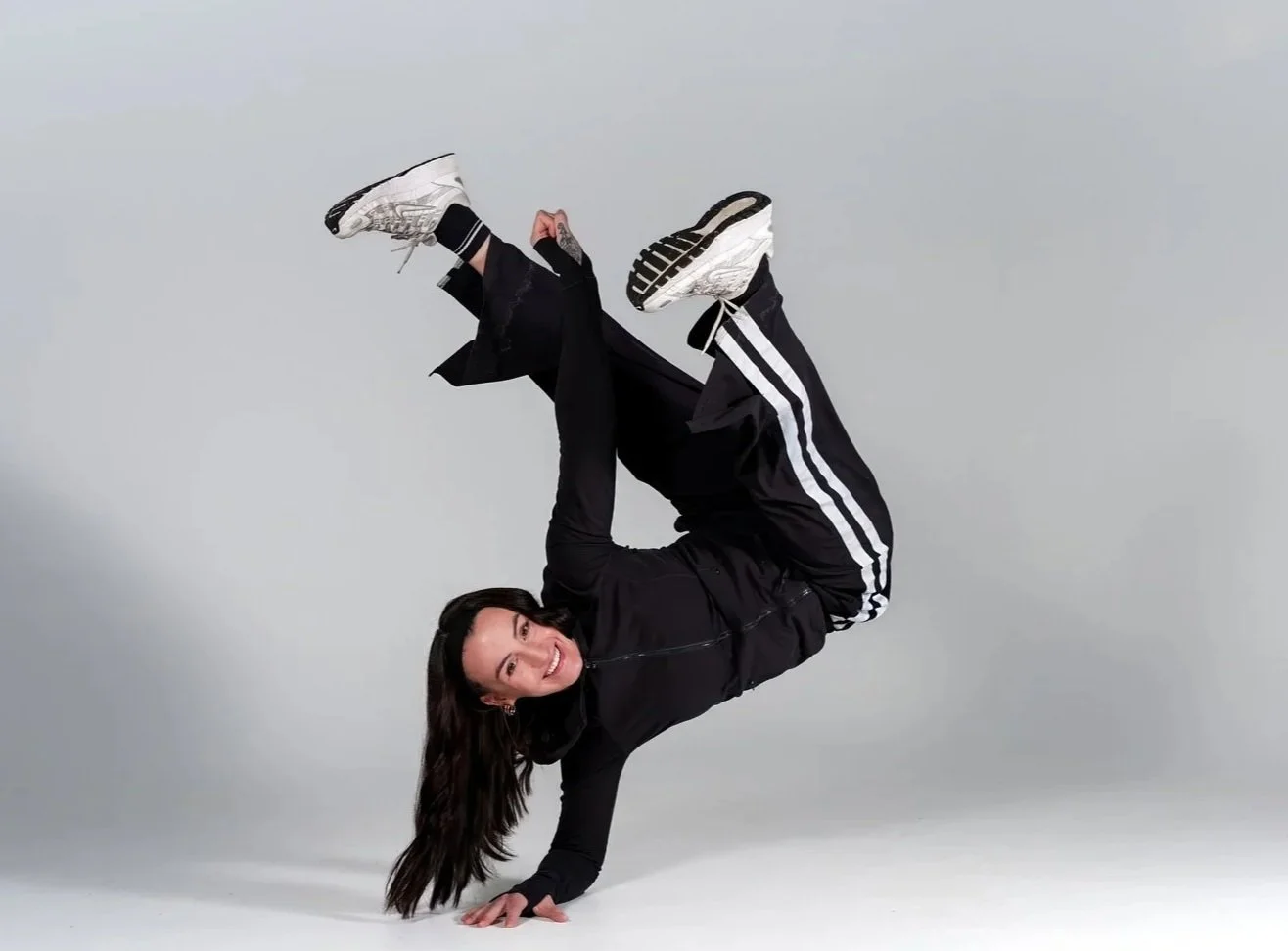 Woman with long dark hair smiling while performing a one-handed handstand, wearing black athletic clothing with white stripes and white sneakers, against a plain light gray background.