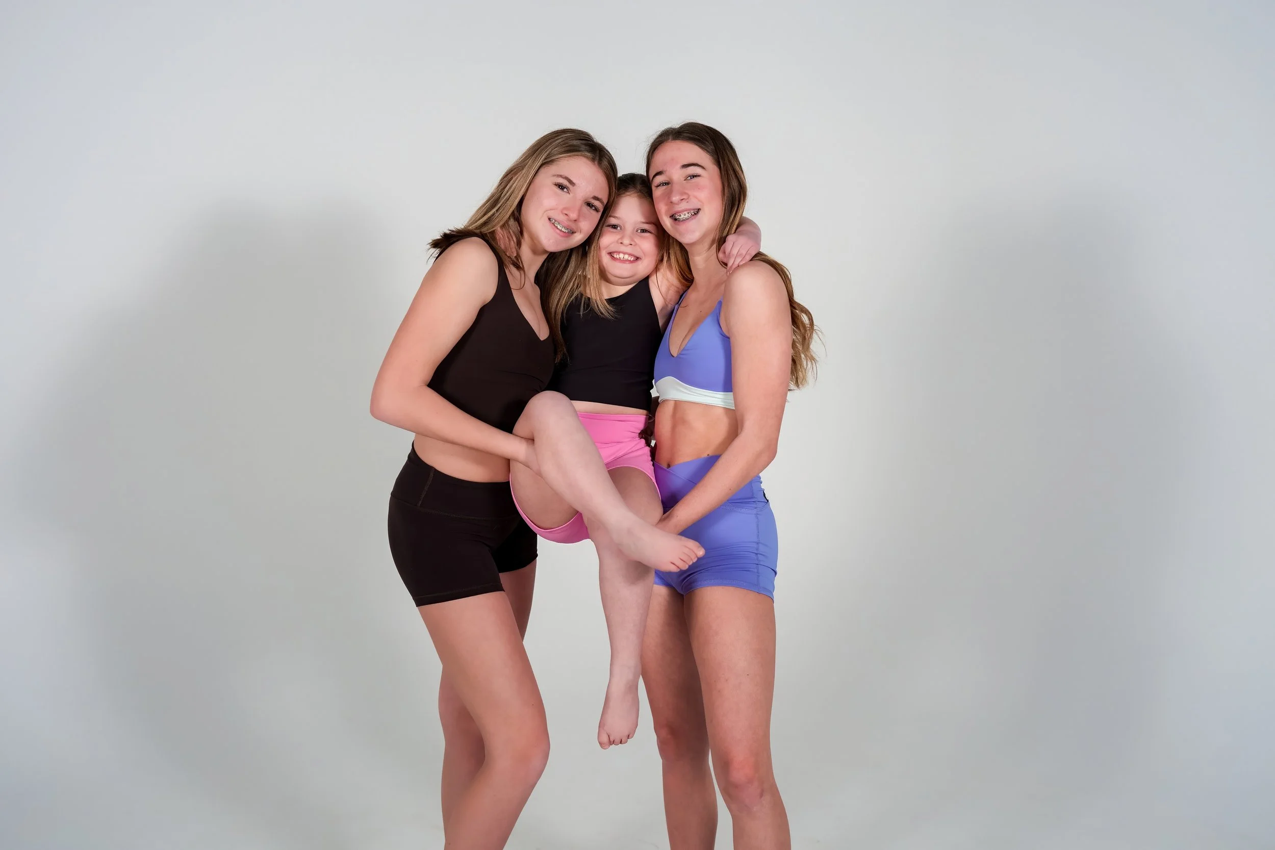 Three girls smiling and hugging each other, with one girl in pink shorts being lifted by the two girls in black and purple sports bras and matching shorts.