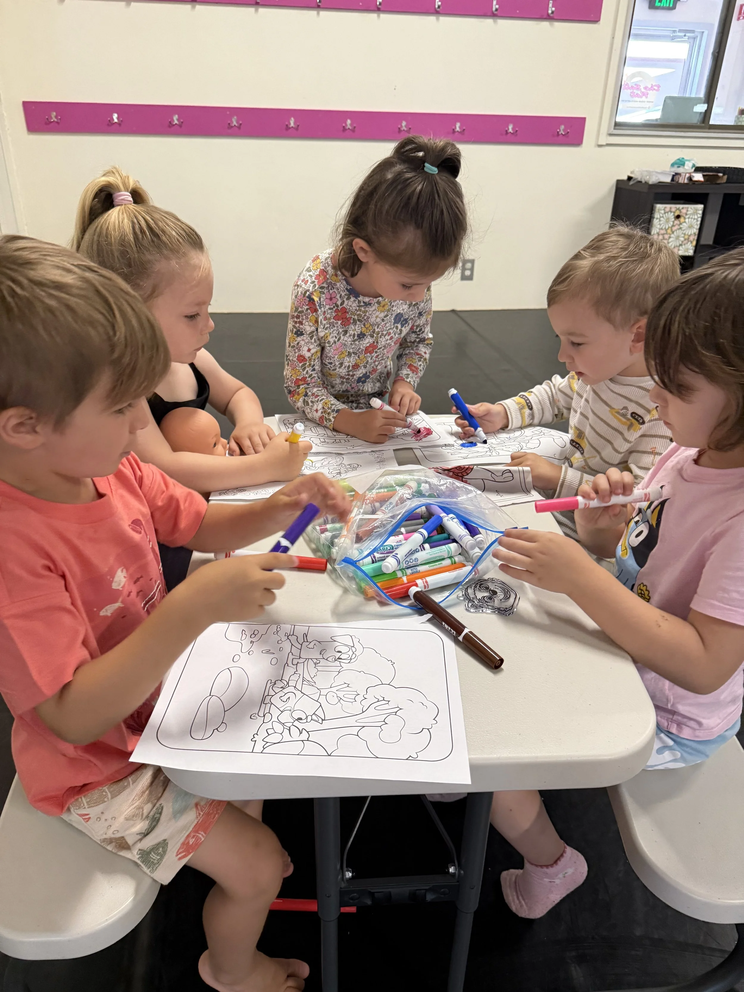 Six children sitting around a table coloring with markers. There are coloring pages, a bag of markers, and a doll on the table. The setting is a classroom with pink hooks on the wall and a window.