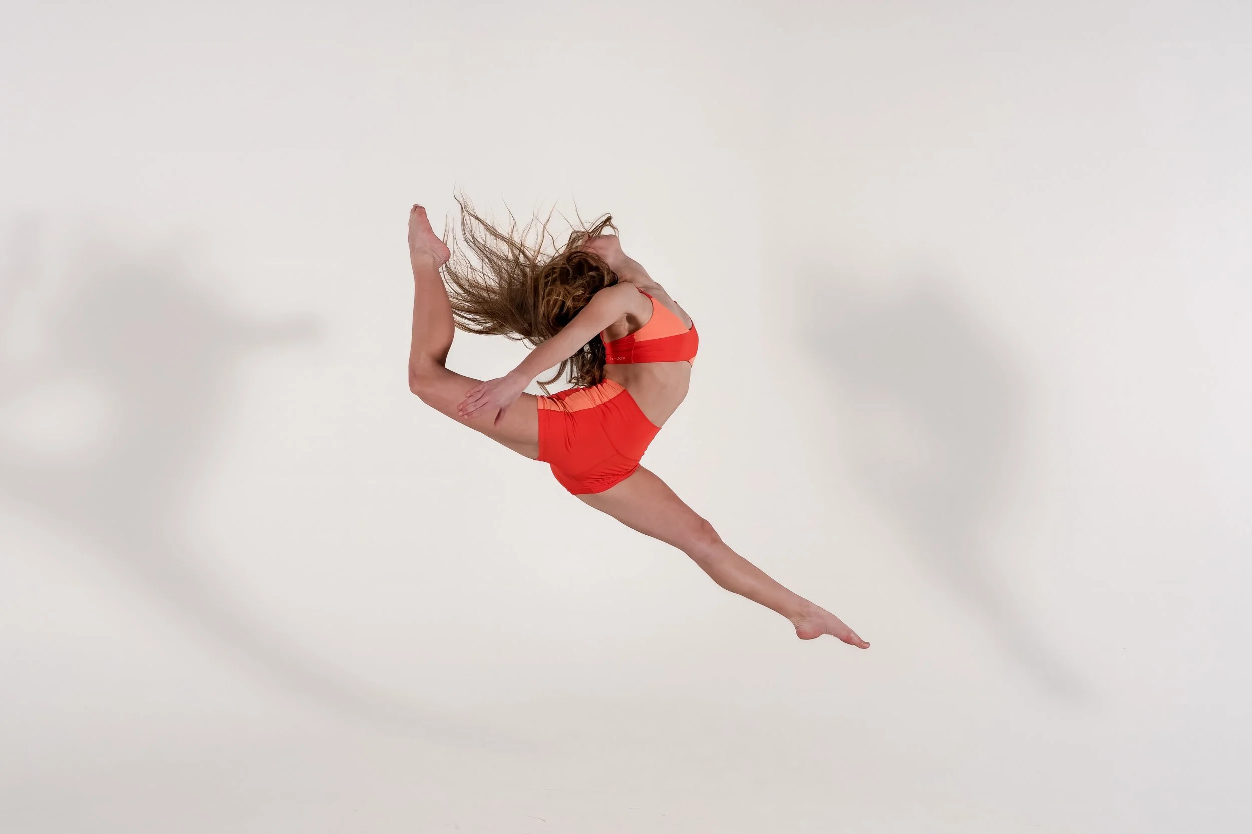 A woman in a leotard performing a contorted dance pose against a plain white background, with long hair flowing and casting a shadow.