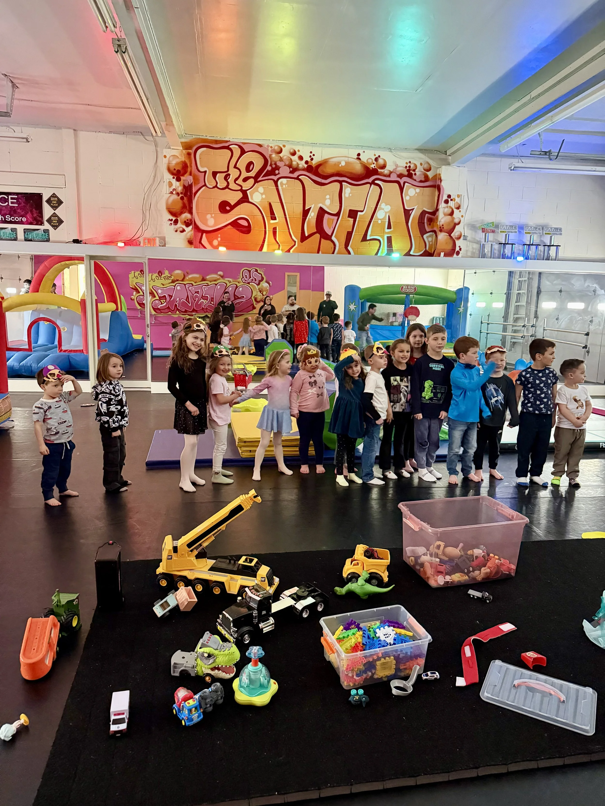 Children standing in line at an indoor play area with colorful inflatables and play equipment, and a large graffiti-style sign that says 'The Salt Flap' on the wall.