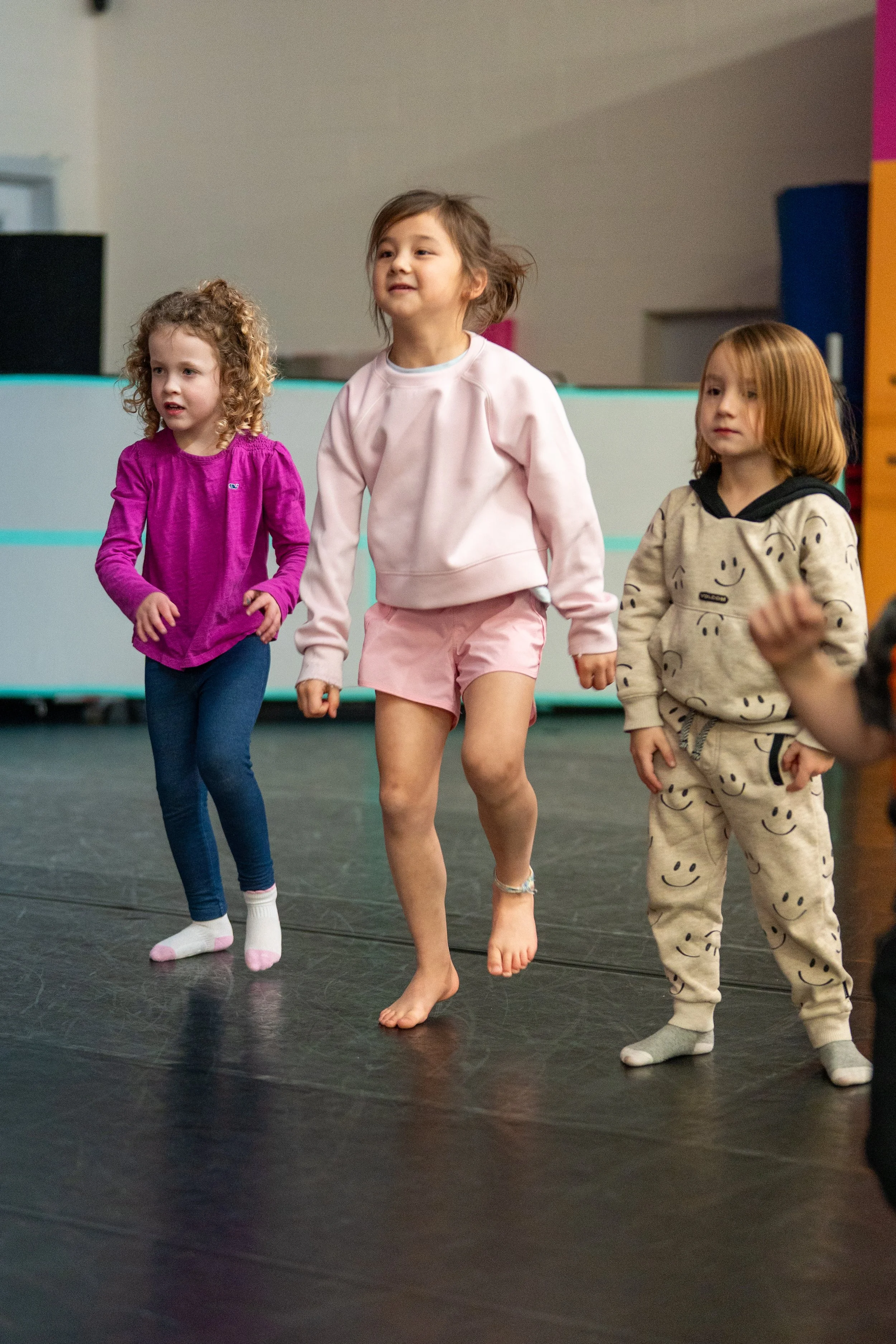 Three young girls are dancing or jumping together indoors with a dark floor and colorful background.