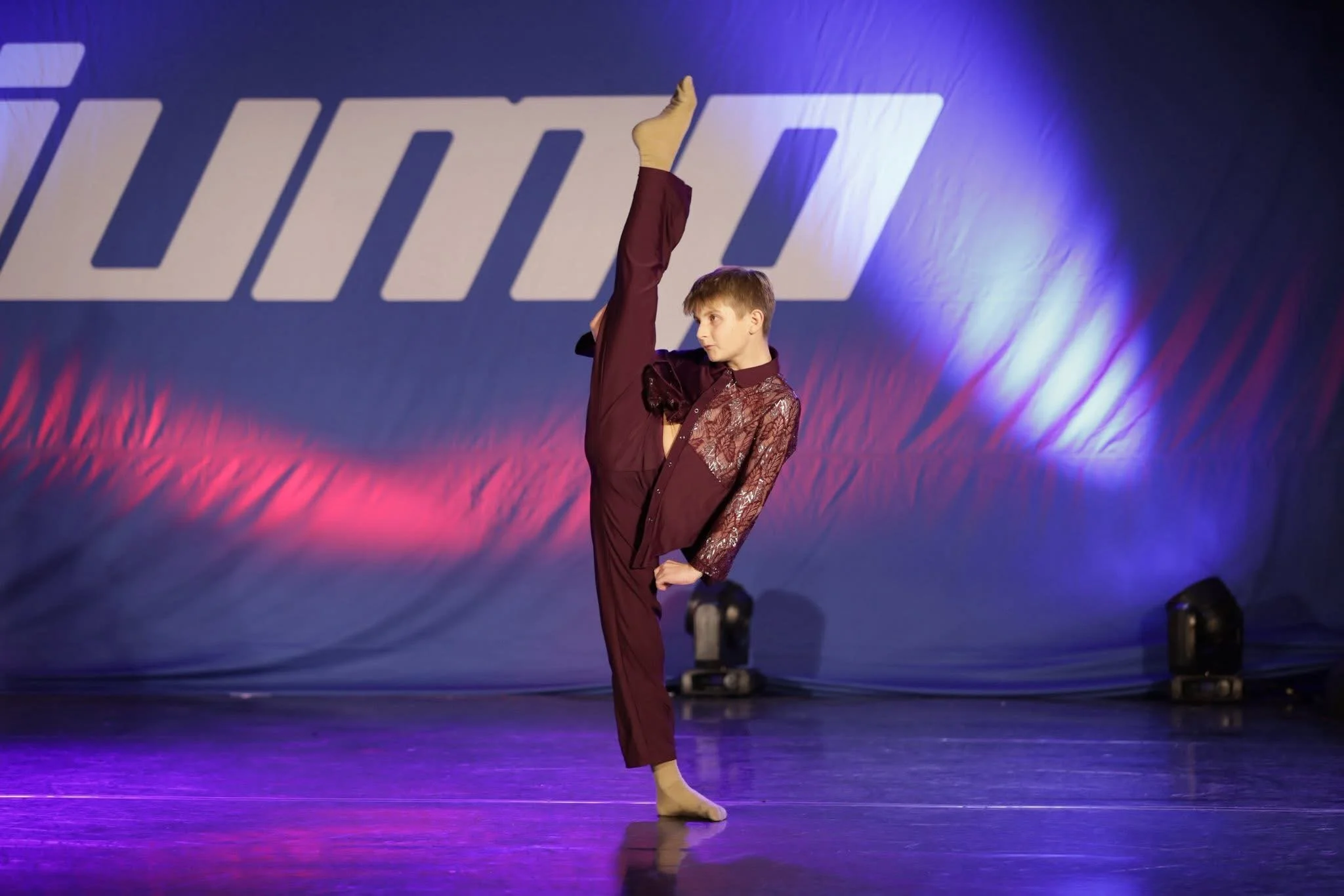 A young male dancer performing a high kick on stage with a large backdrop featuring stylized text. Stage lighting in purple and pink hues.