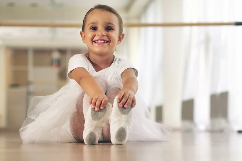 Young girl in a white dress and socks sitting on the floor in a dance studio, smiling at the camera.