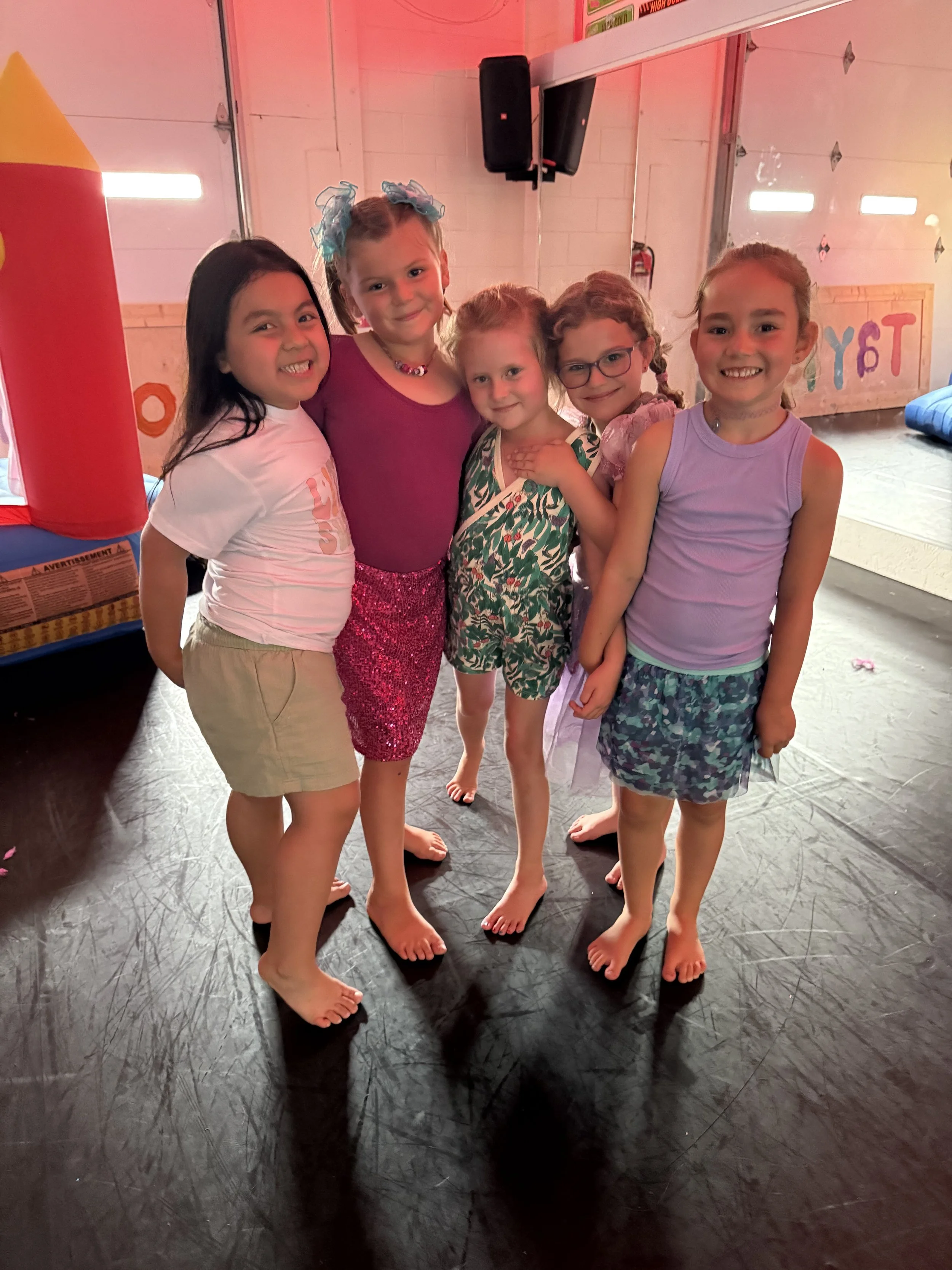 Six young girls standing together on a dance studio floor, smiling at the camera. They are barefoot and dressed in colorful casual clothing, with some wearing skirts and others shorts. The background features a birthday party setup with a bounce house and a decorated wall.