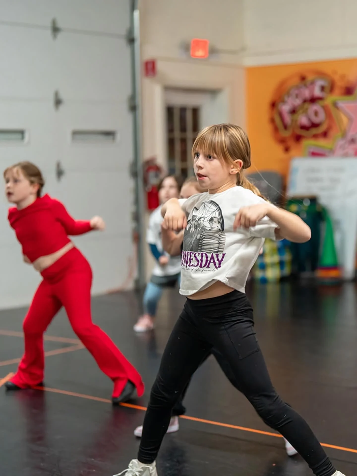Children dancing in a dance class, with a girl in a white T-shirt and black pants in the foreground, and a girl in a red outfit in the background, inside a studio with colorful graffiti on the wall.