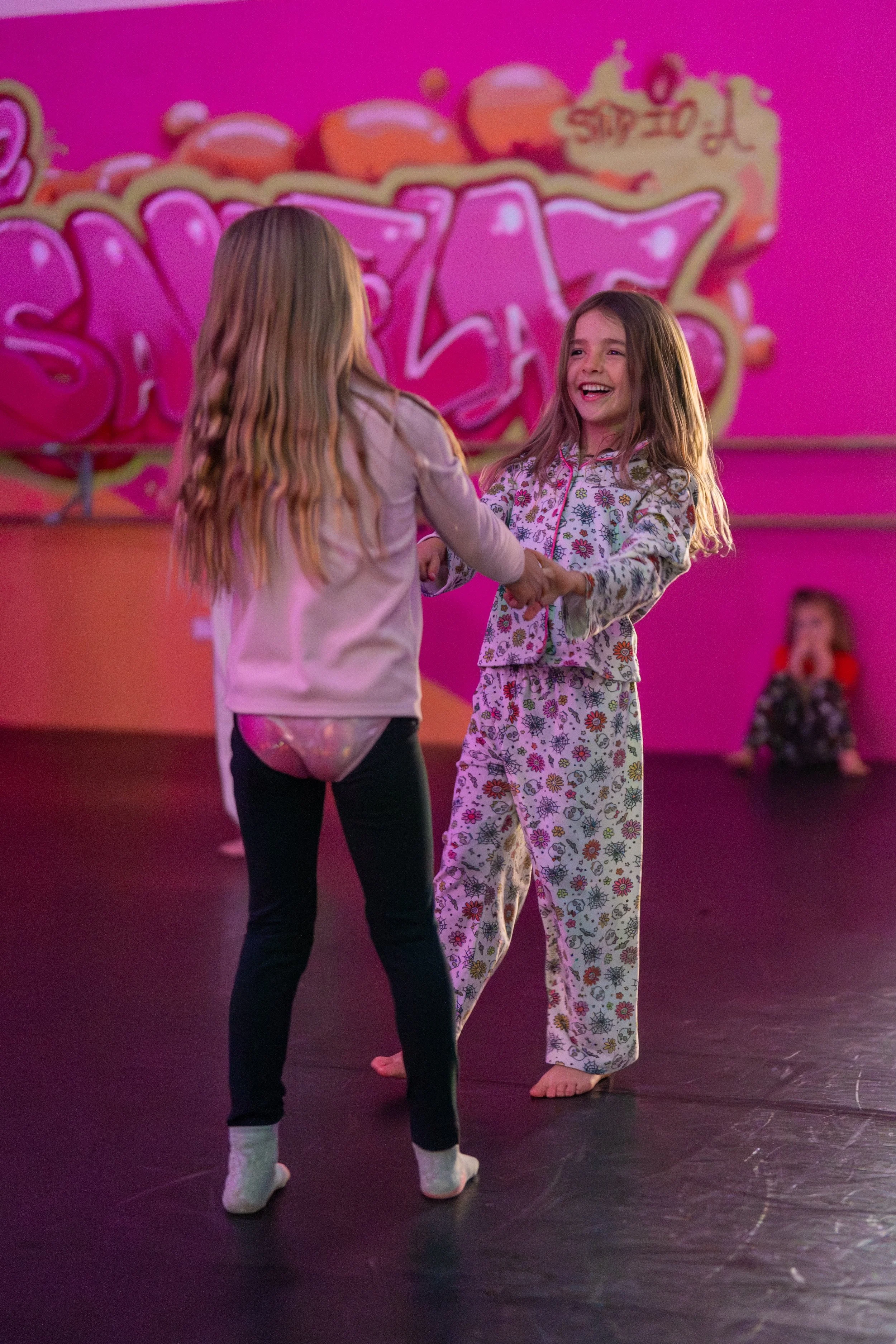Two young girls holding hands and smiling at each other during a sleepover in pajamas, with another girl sitting in the background at a pink-themed party.