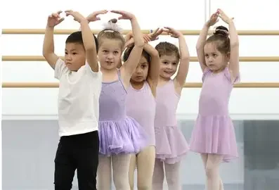 Five young children in ballet outfits standing at a ballet barre, with the boys and girls raising their arms above their heads.