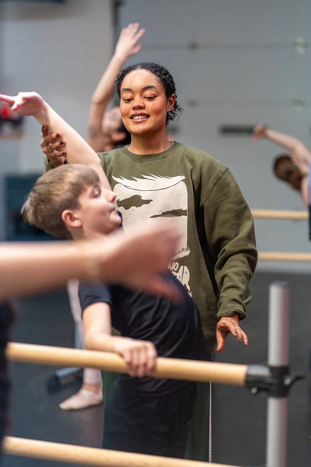 A ballet instructor, a young woman with curly black hair, is smiling while instructing children in a dance class at a barre. One young boy in black is holding the barre and looking at her, while other children are stretching their arms in the background.
