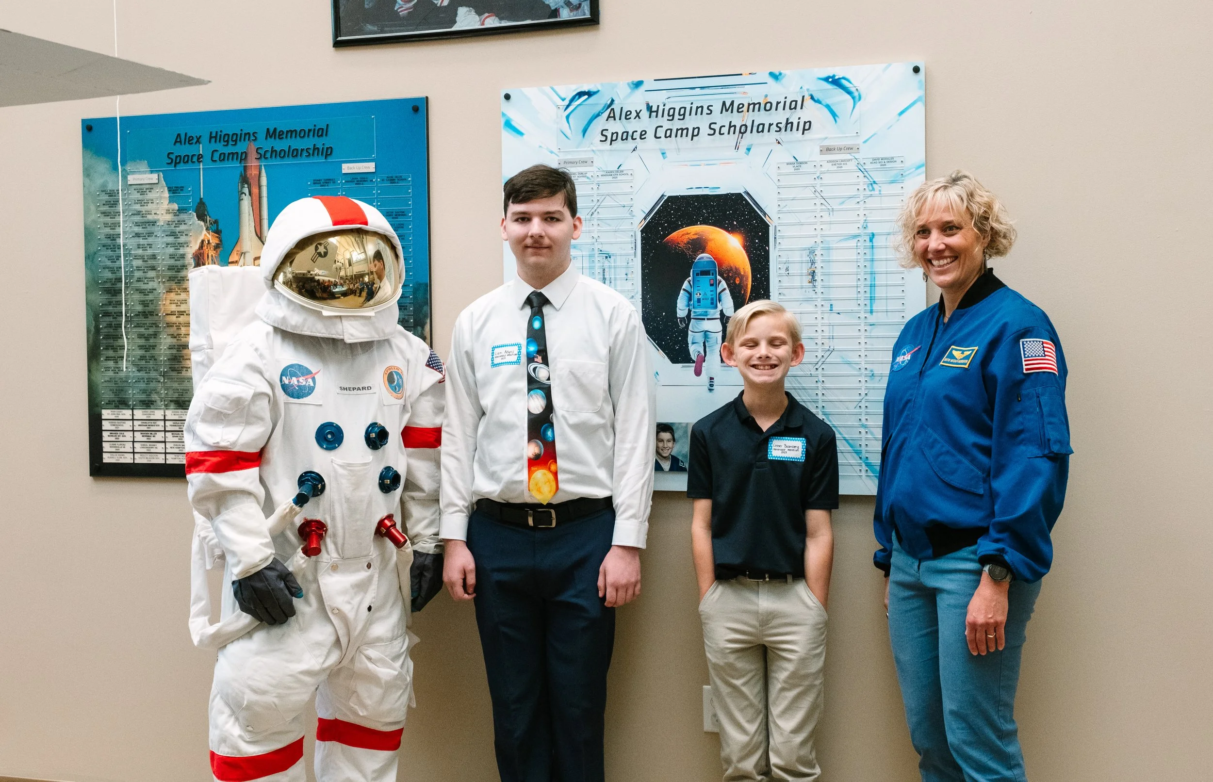 Four people standing in front of space-themed posters, including an astronaut in a white spacesuit with NASA patches, a young man in a dress shirt and tie, a young boy in a black shirt, and a woman in a blue NASA jacket, smiling at the camera.