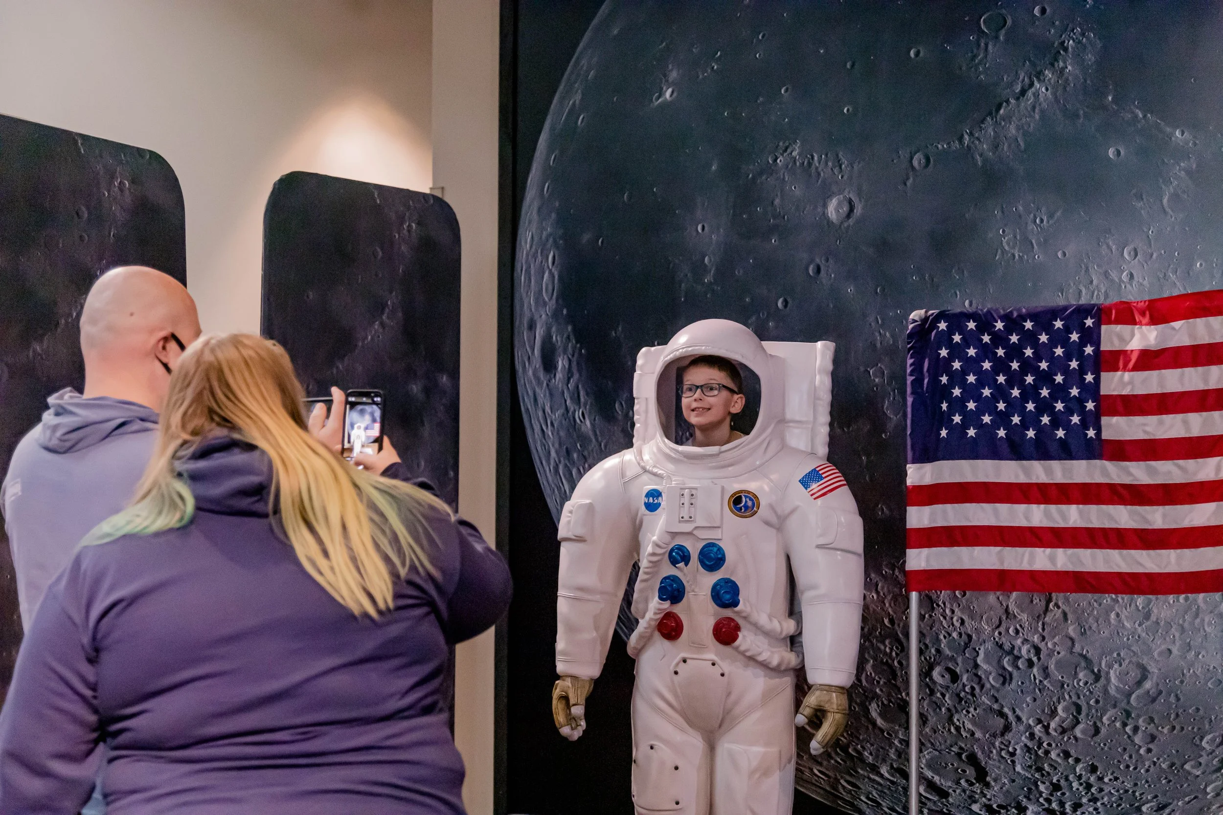 People taking photos of a boy in an astronaut suit at an exhibition, with lunar and American flag backgrounds.