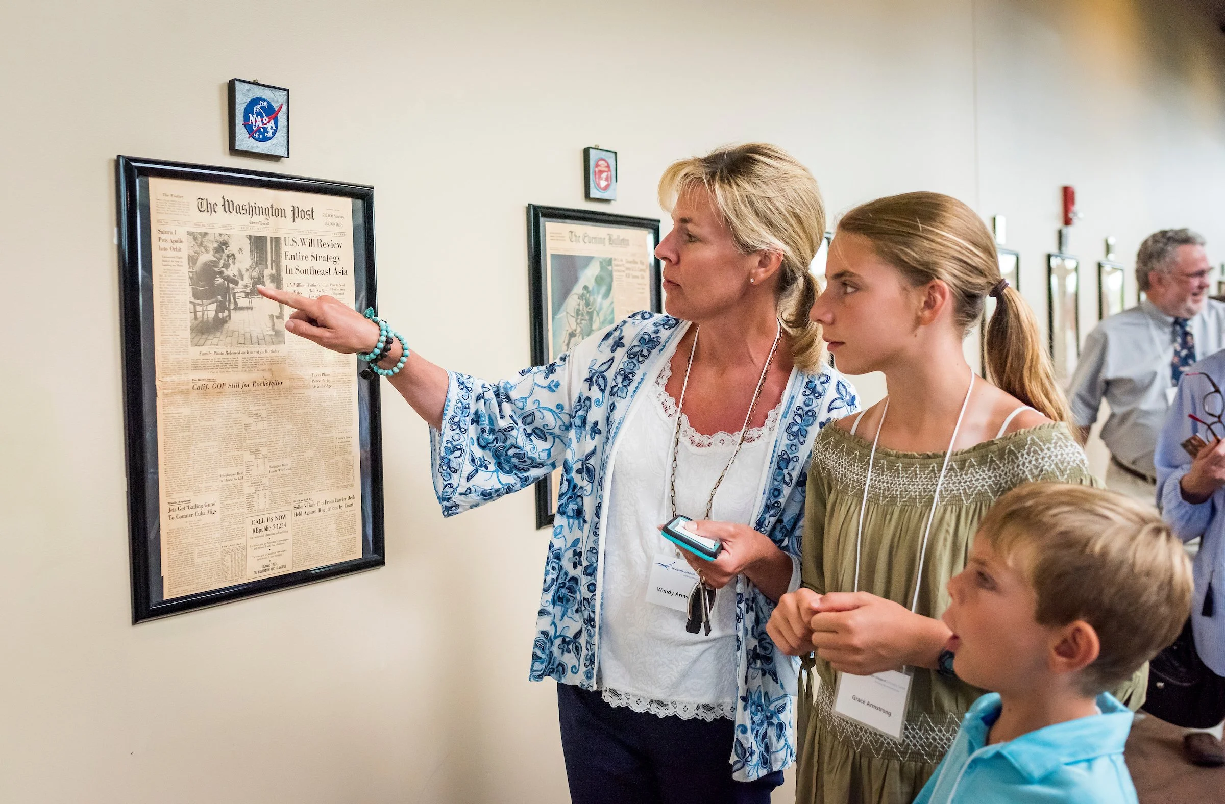 Two women and several children look at framed newspaper articles and NASA patches on a wall in an indoor space. The woman on the left, with short blonde hair, is pointing at a newspaper, and the girl beside her has pulled her hair back into a ponytail.