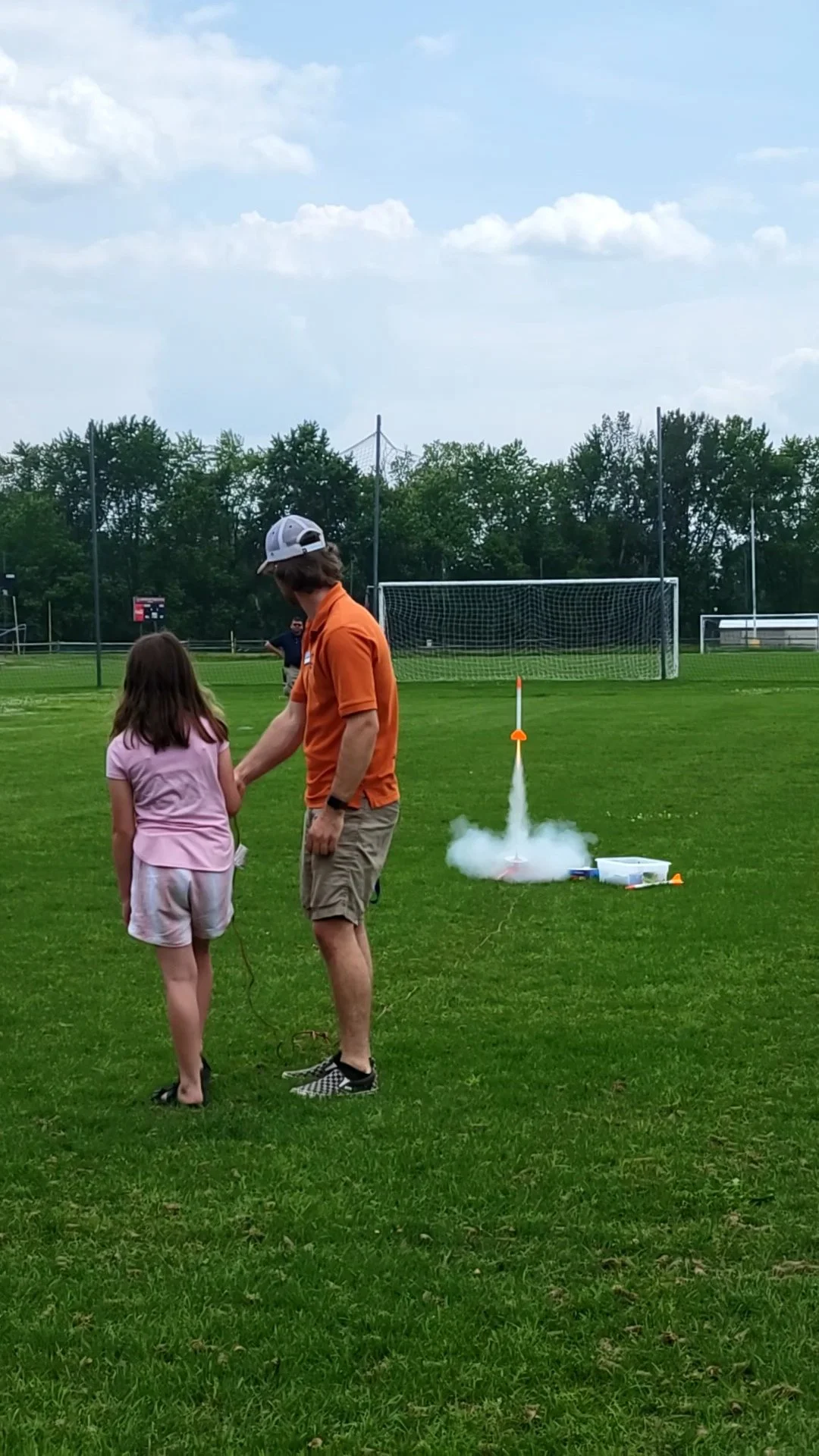 A man and young girl hold hands on a grassy field near a model rocket that is launching, with smoke coming out of the rocket. The girl is wearing a pink shirt and shorts, and the man is dressed in an orange shirt, khaki shorts, and a white baseball cap. In the background, there are trees, a soccer goal, and a cloudy sky.