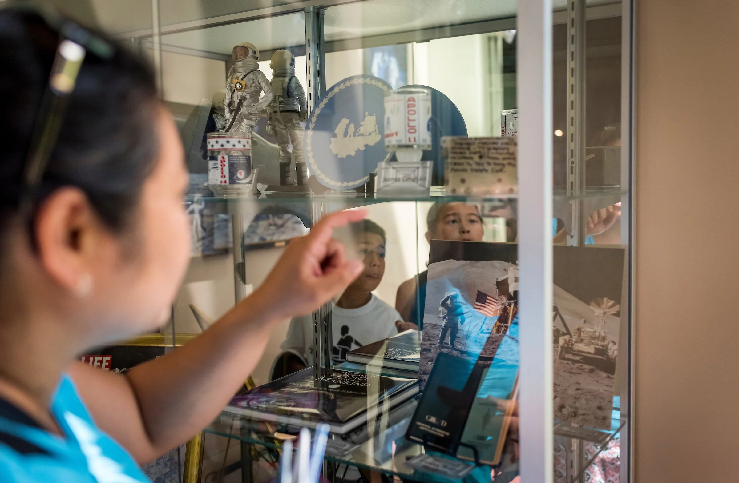 Young girl with glasses looking at a display case containing astronaut figurines, books, and photographs related to space exploration.