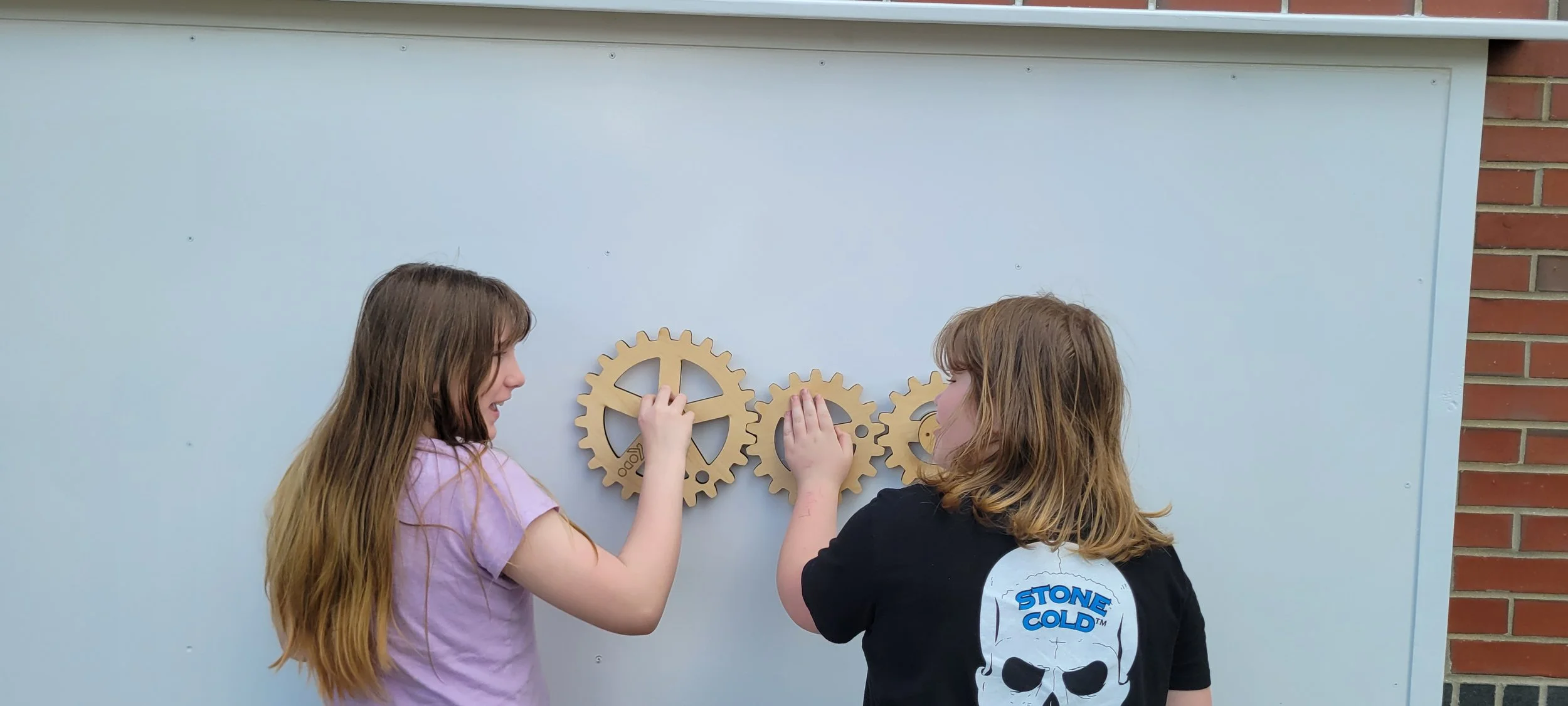 Two girls with long hair, facing each other and touching large wooden gears attached to a blue wall, with a brick wall on the right side.