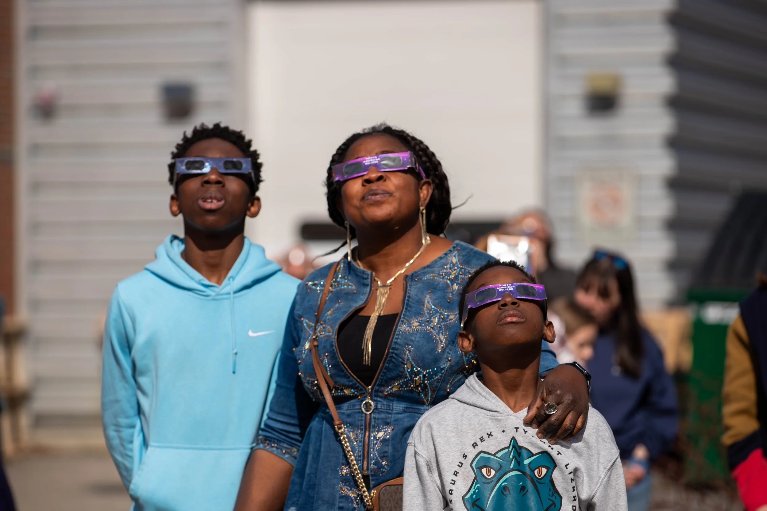 A diverse group of people, including adults and children, wearing eclipse glasses while watching a solar eclipse outdoors. They are standing close together in front of a staircase and a building with white siding.