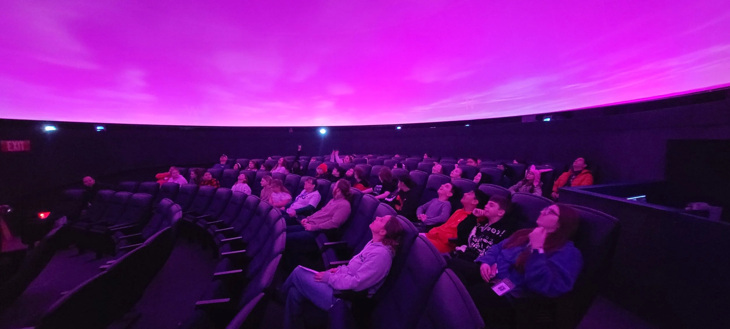 People seated in a theatre watching a film on a large curved screen with a purple sky display.