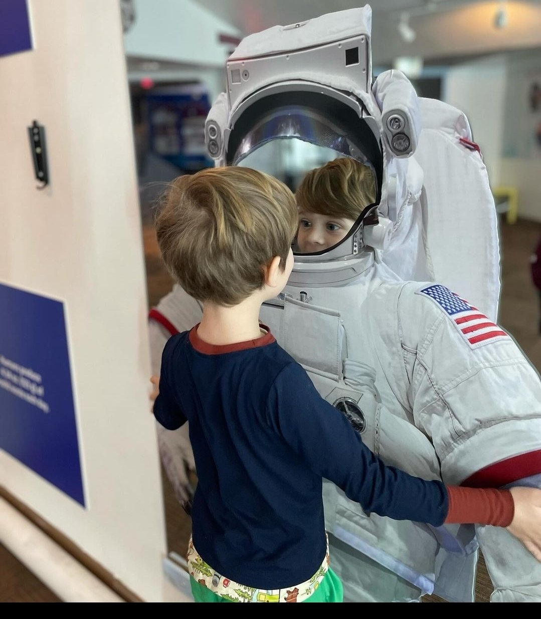 A young boy with brown hair and a blue shirt meets an astronaut in a white space suit with an American flag patch. They are indoors, with the boy touching the astronaut's arm and looking at the astronaut's reflected face in the helmet's visor.