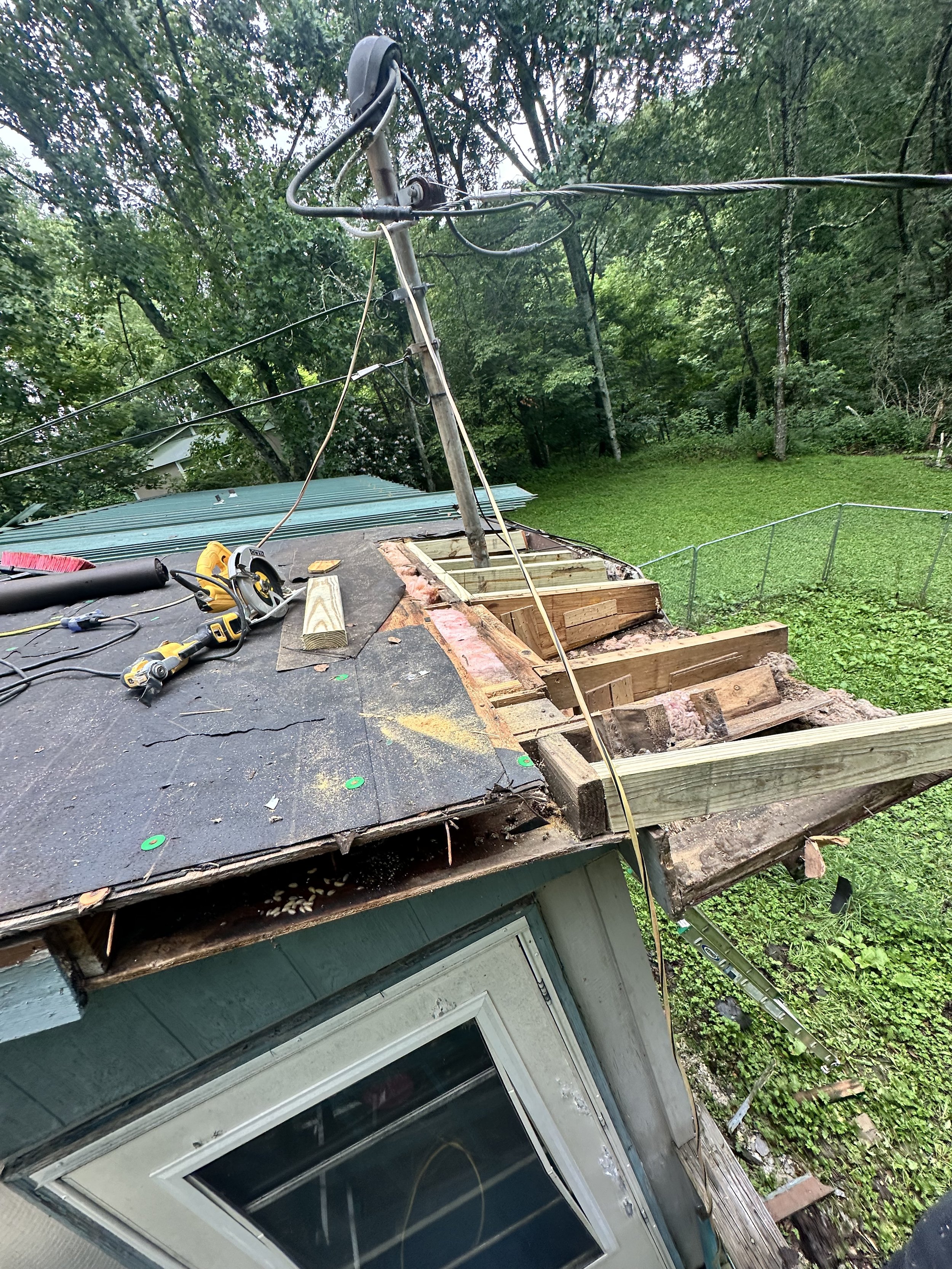 The image shows a house with a damaged roof, missing shingles, and exposed wooden beams. There are tools and construction materials on the roof, and a damaged power pole leaning to the side with wires hanging loose. The house is surrounded by green trees and grass.