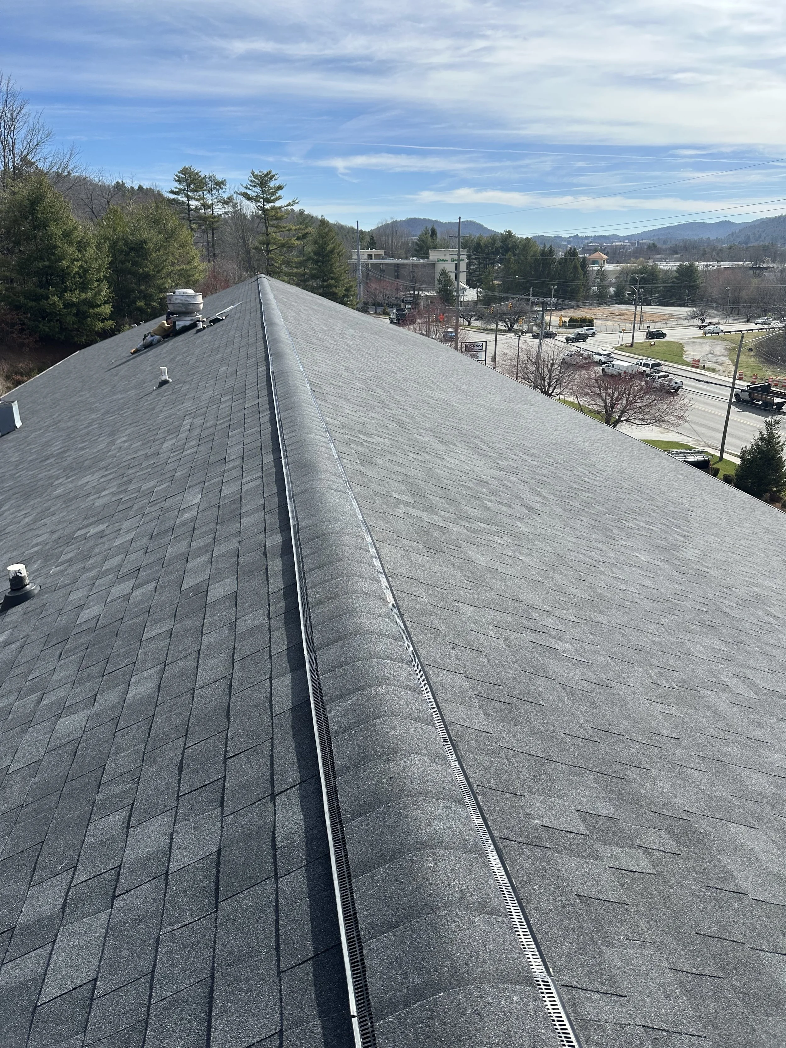 A rooftop with gray shingles and a metal ridge vent, overlooking a suburban area with trees, streets, and a partly cloudy sky.