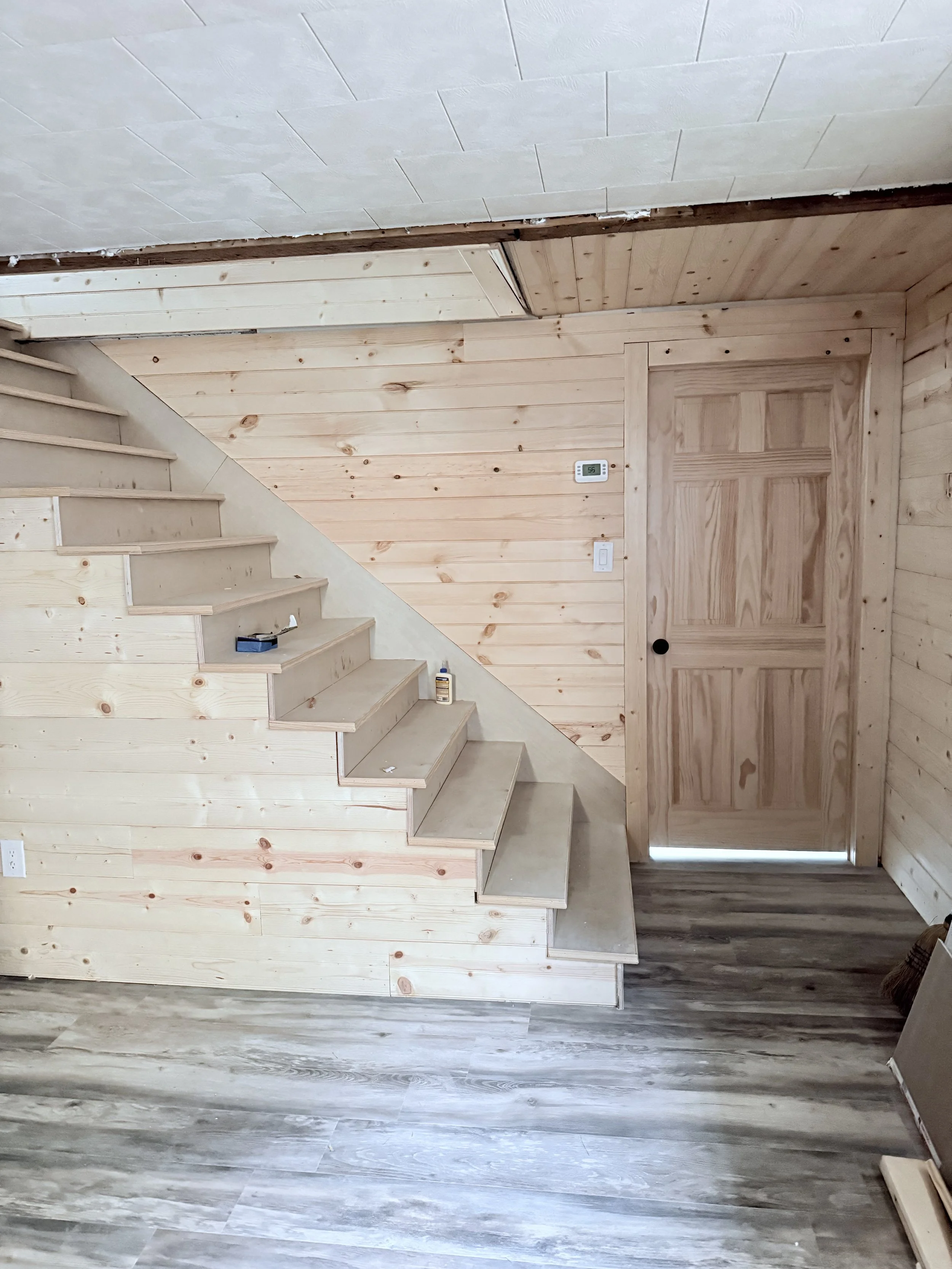 Interior of a wooden staircase with light-colored wood paneling on the walls, a closed wooden door, a thermostat on the wall, and a section of how to finish the staircase with new wood steps.